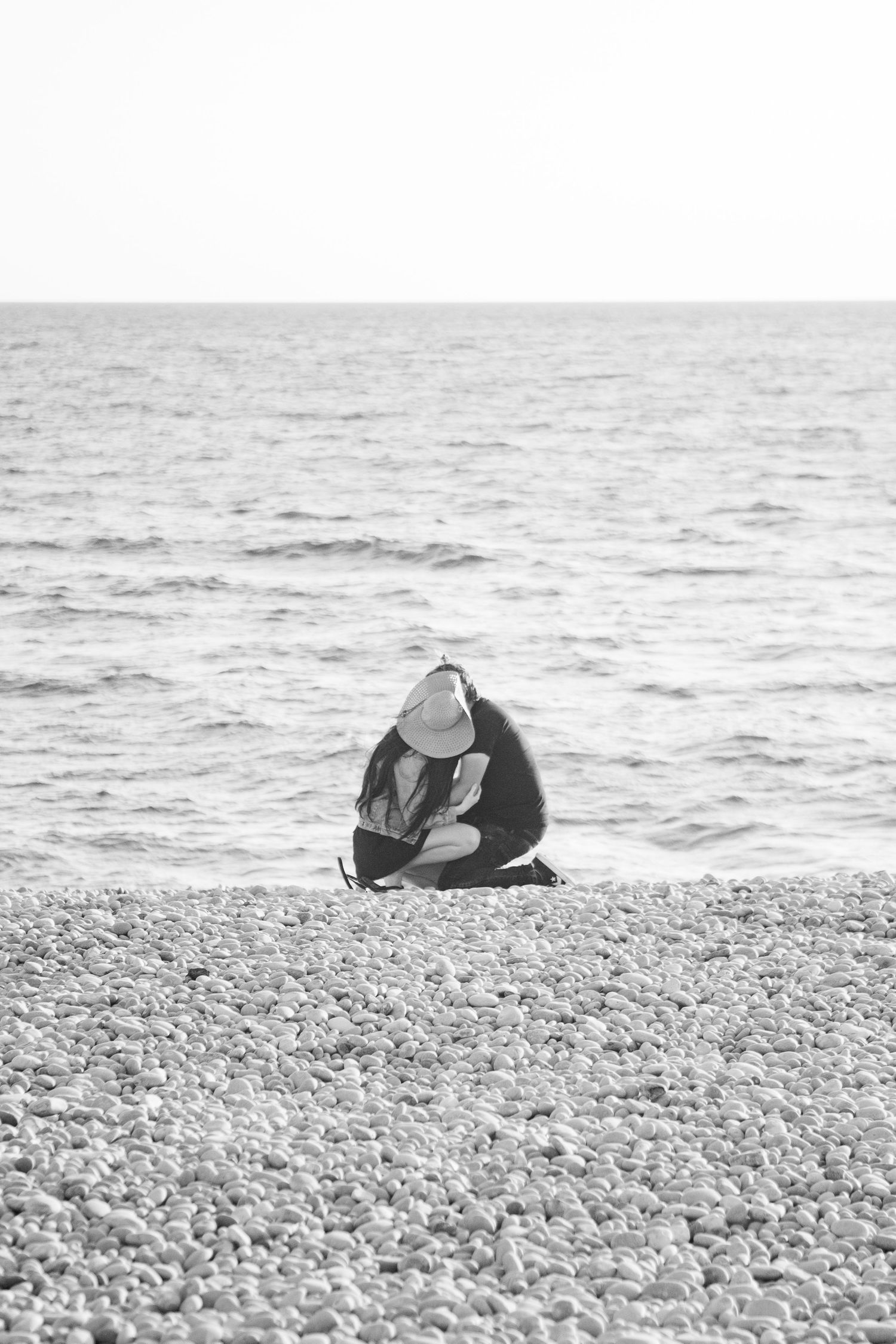 Black and white photo of a couple knelt down on the beach, wrapped in an embrace in the distance. Stony beach in front, and the sea behind.