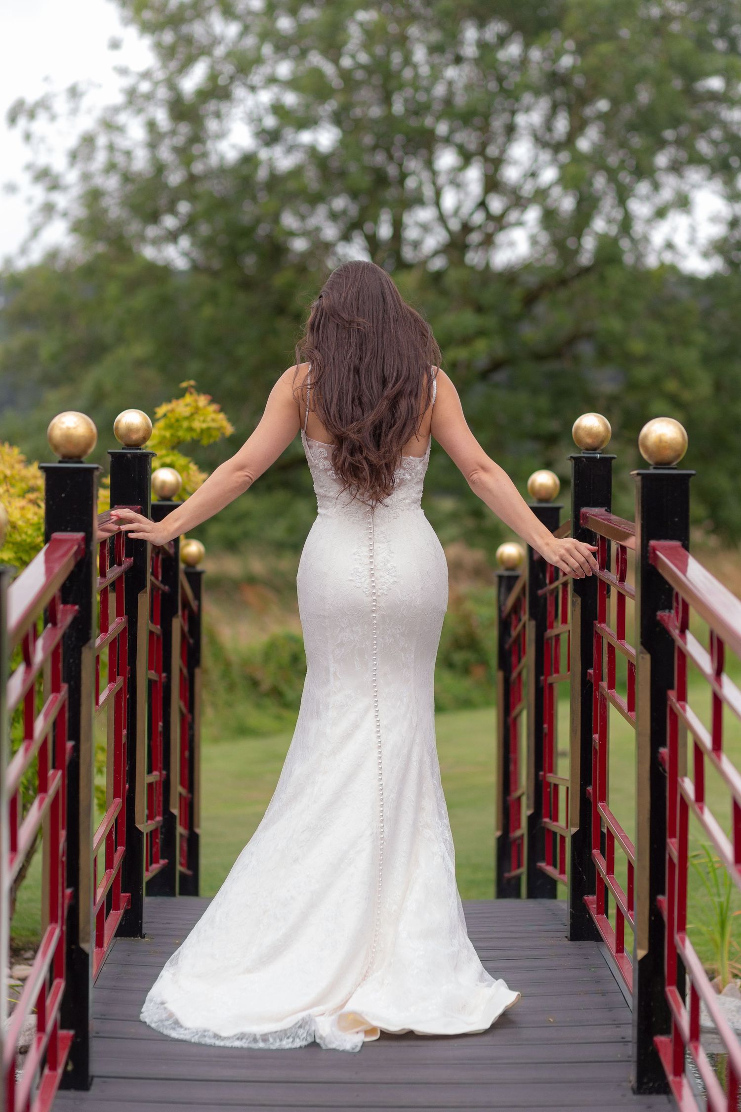 A bride walks away from the camera across a black and red bridge, arms stretched out for the hand rails, buttons flowing down her back