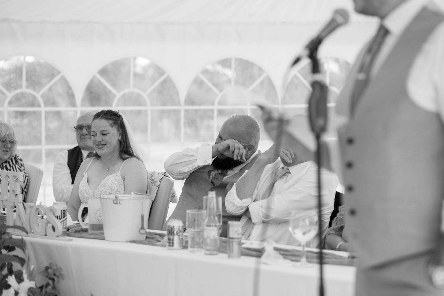 A wedding top table, with the Bride laughing and the Groom wiping away happy tears as his Best Men give a speech.