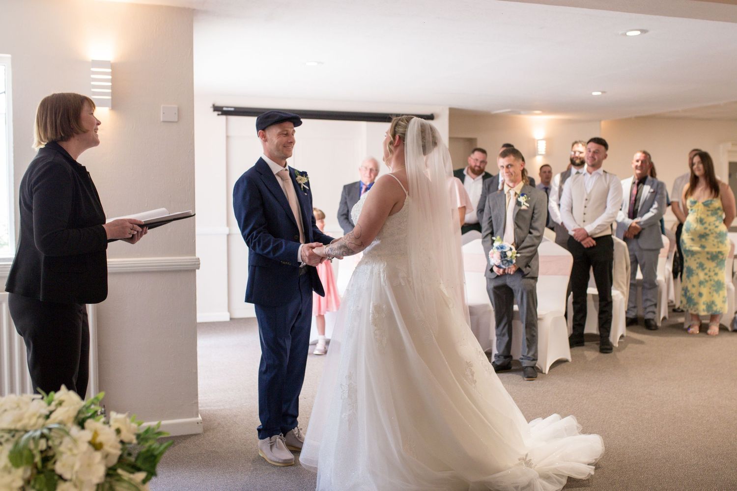 Bride and Groom stand hand in hand as the registrar talks them through their wedding ceremony. A room full of guests stood behind them