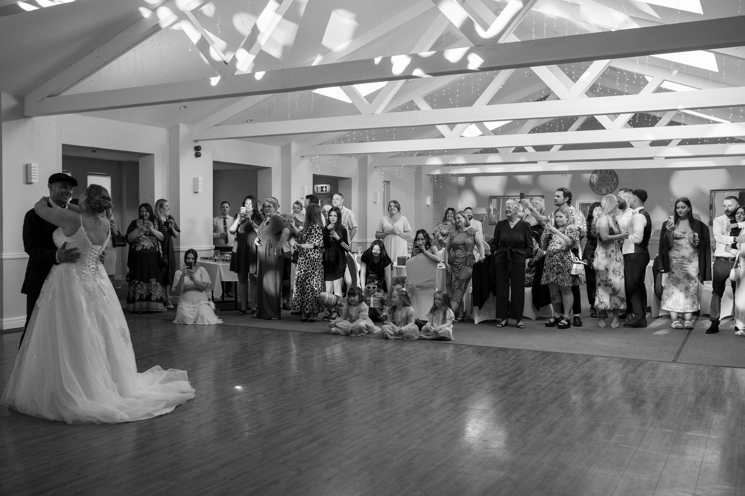 Black and White photo of a bride and groom sharing their first dance, with their guests watching on behind them , `