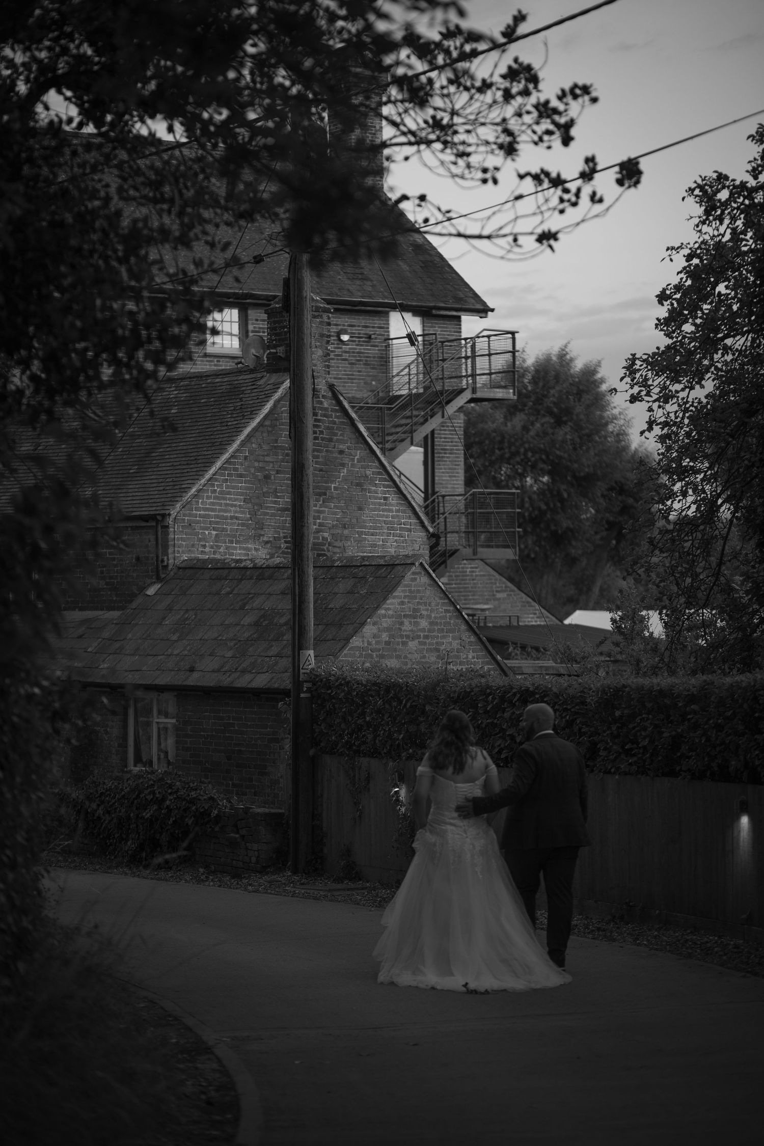 Black and white photo of bride and groom walking back towards Sopley Mill, with their arms wrapped round each other.