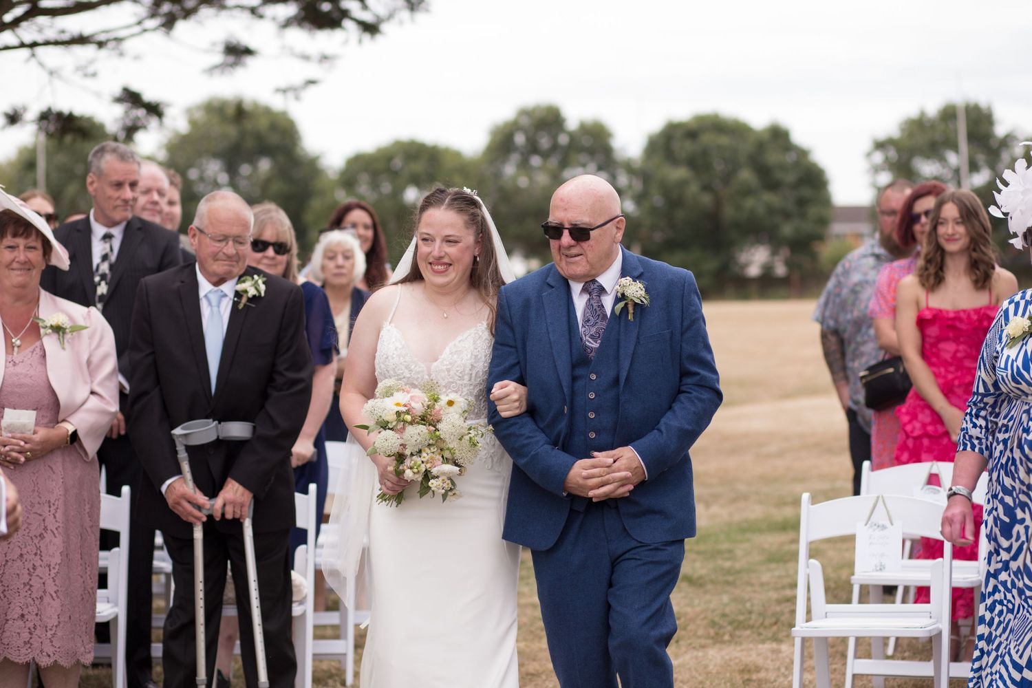 Bride and her father walk down the aisle, surrounded by their guests, during an outdoor ceremony at Weymouth and Portland Rugby Club