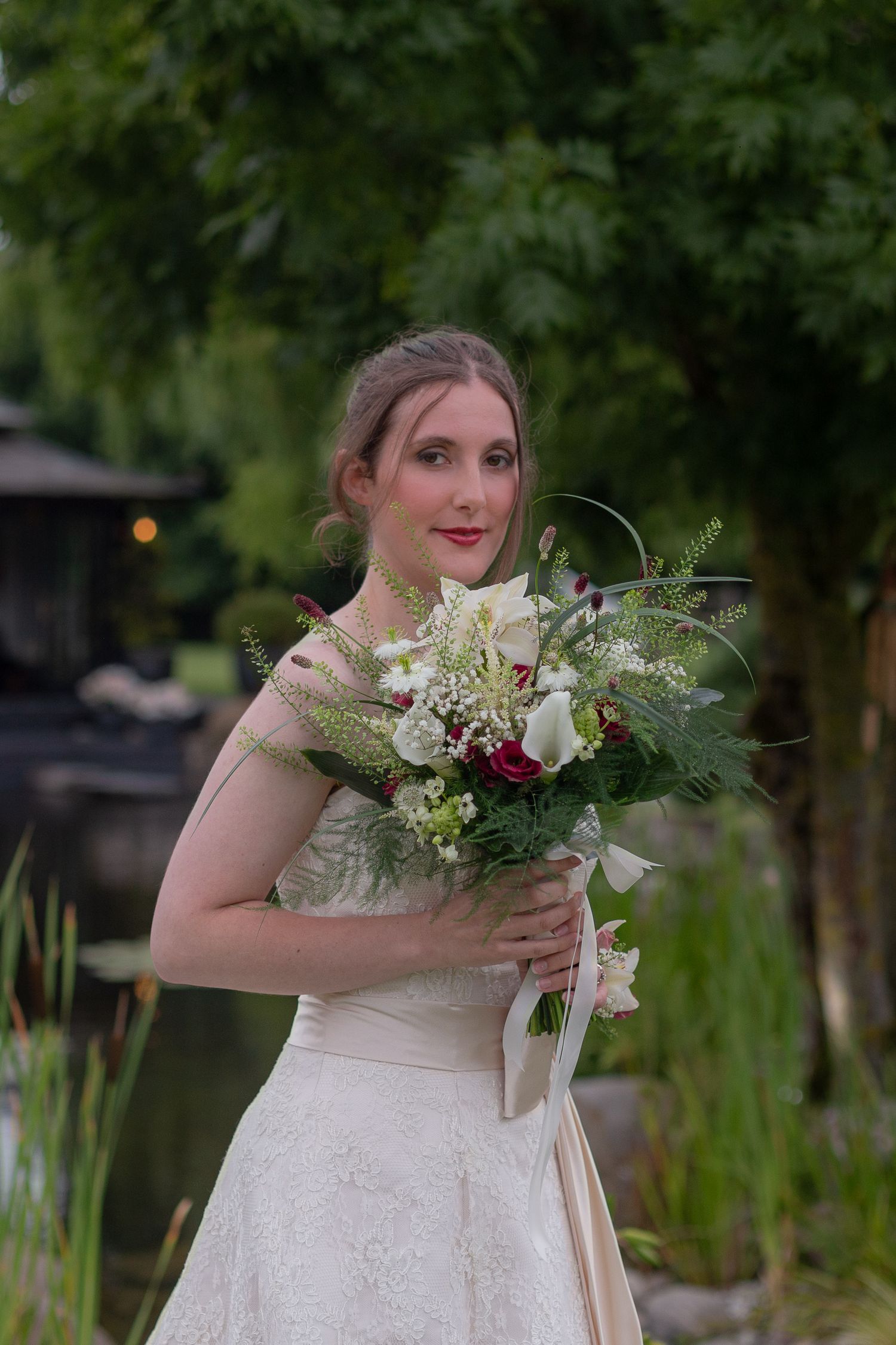 A bride poses for a portrait, holding up a beautiful big bouquet of white and red flowers with lots of greenery.