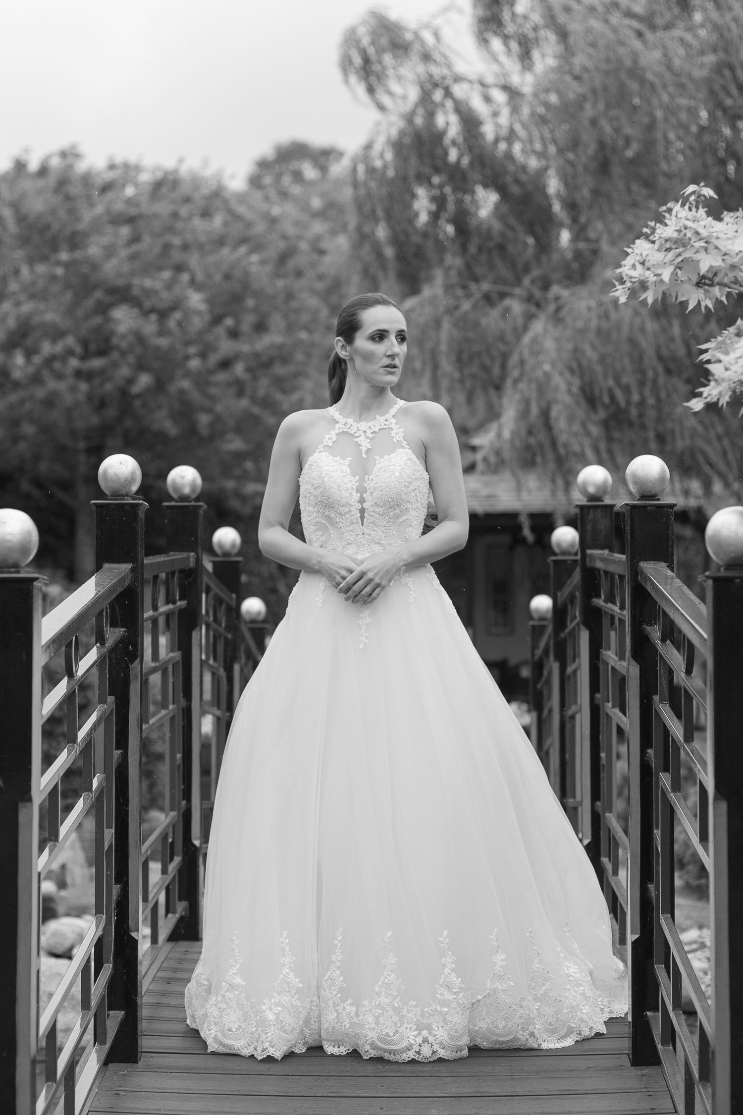 A bride stands in the middle of a bridge in a ballgown, hands folded gracefully together as she looks off into the distance.