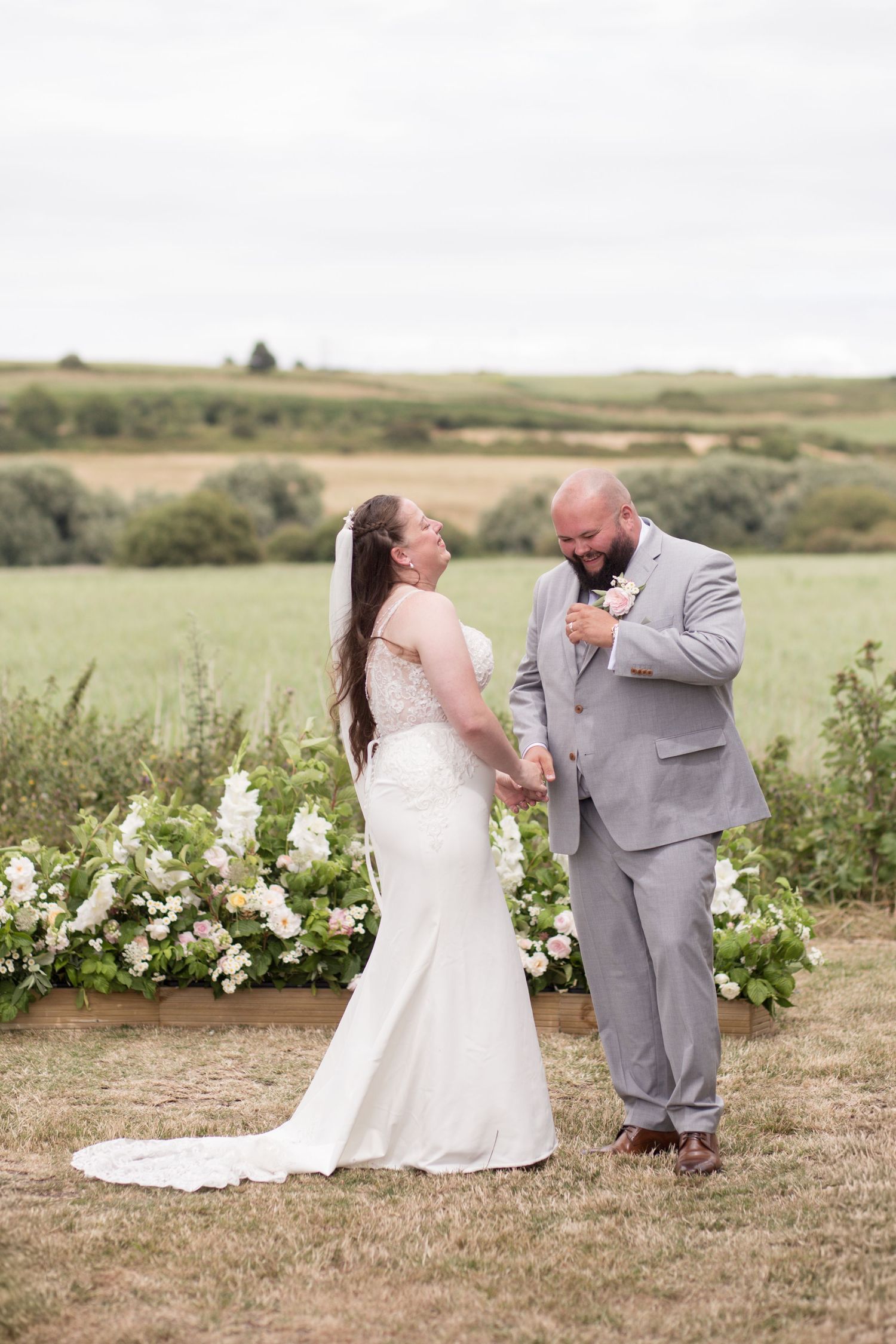 Bride and Groom laugh together in the middle of their outdoor ceremony at Weymouth Rugby Club, with green hills and fields in the background