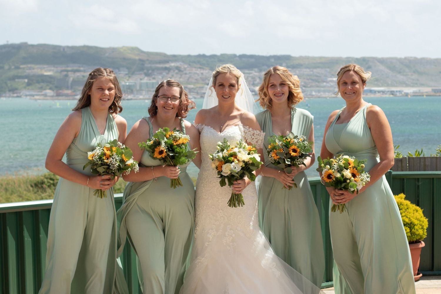 Bride and her Bridesmaids pose for a group photo. Bridesmaids wearing sage green, and all holding bouquets of sunflowers.