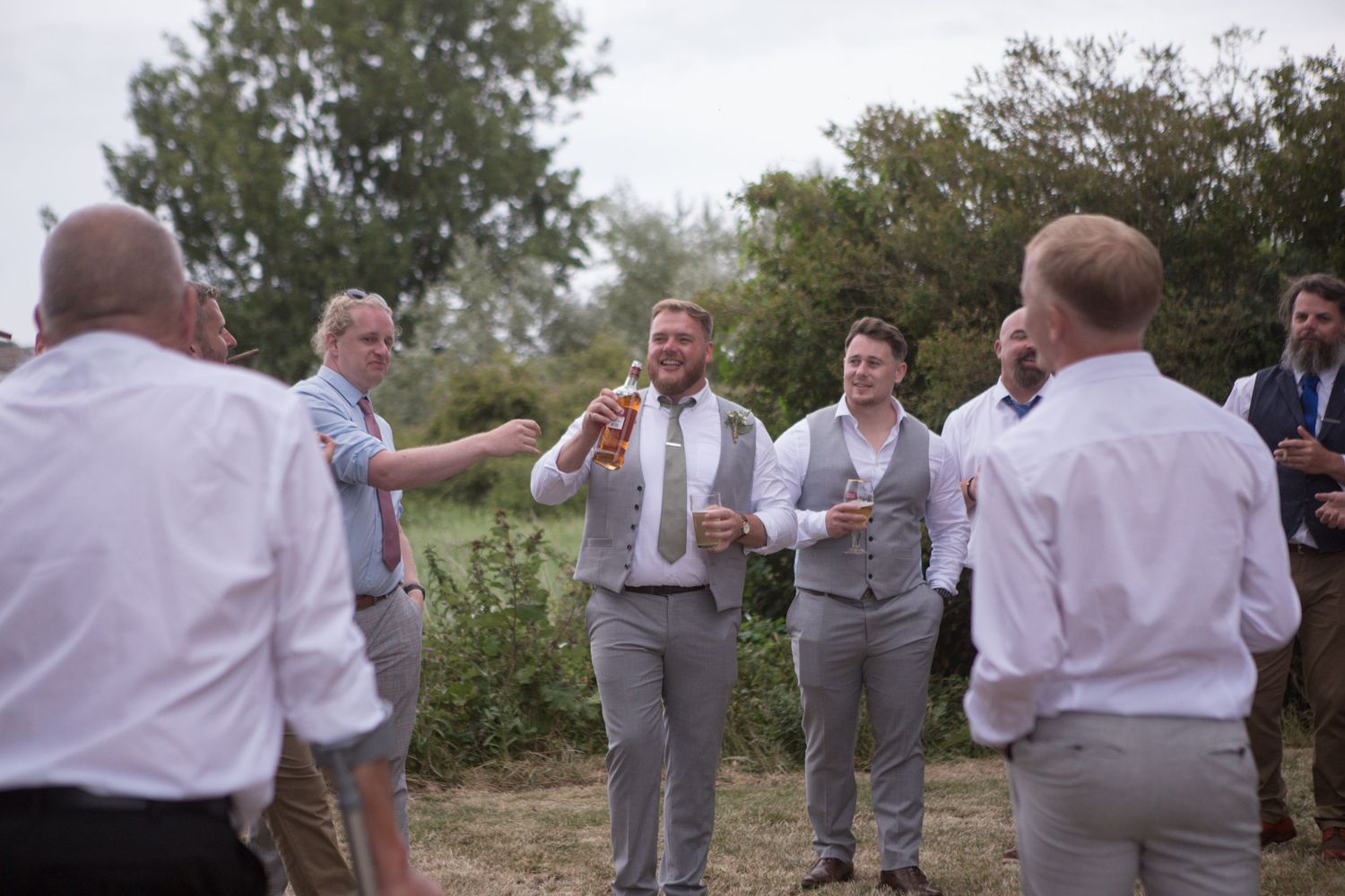 Groomsmen and male wedding guests stand in a circle smoking cigars and passing a bottle of whiskey around between them all.