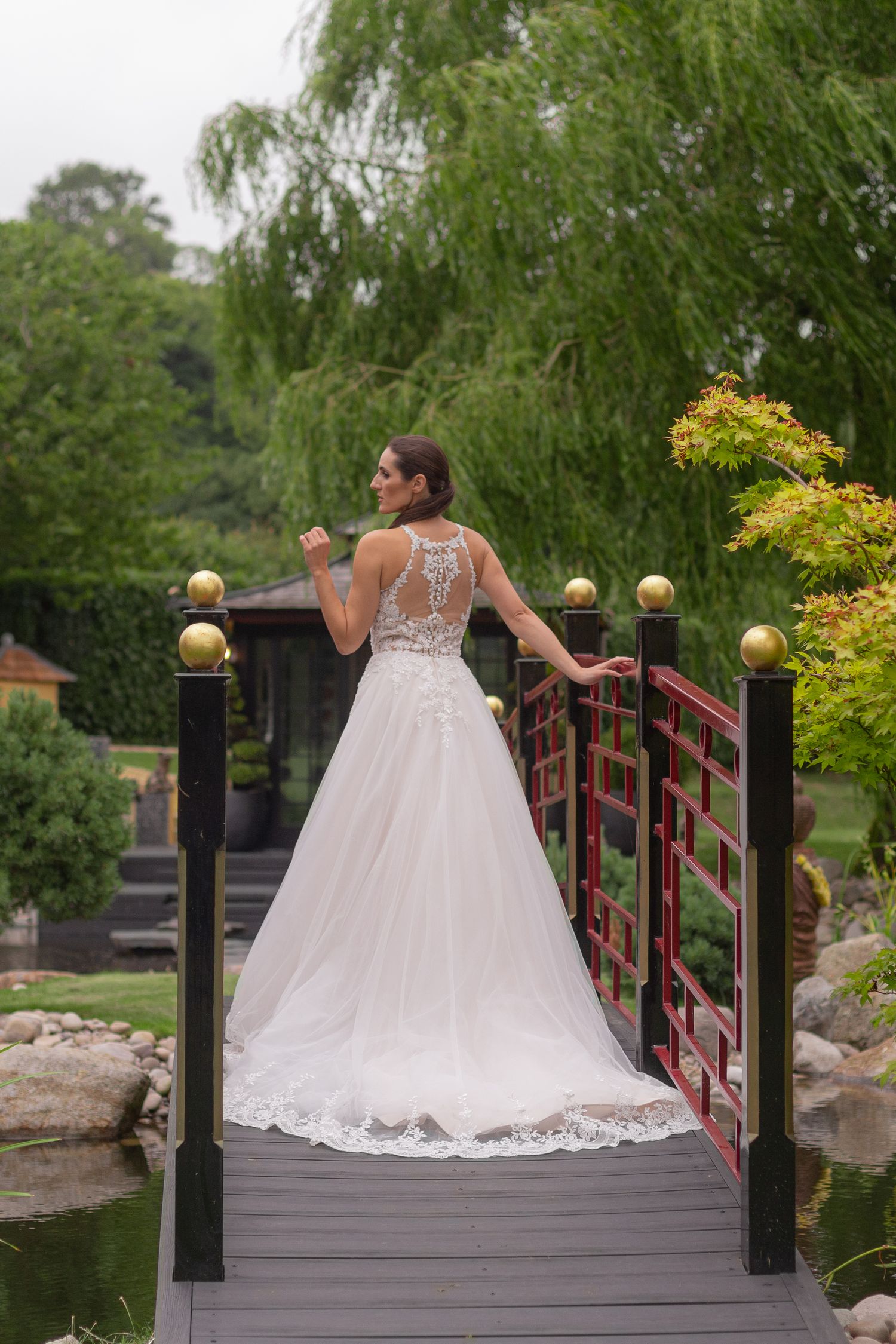 A bride poses for a photo on a little bridge. She faces away from the camera, showing off the back of her ball gown.