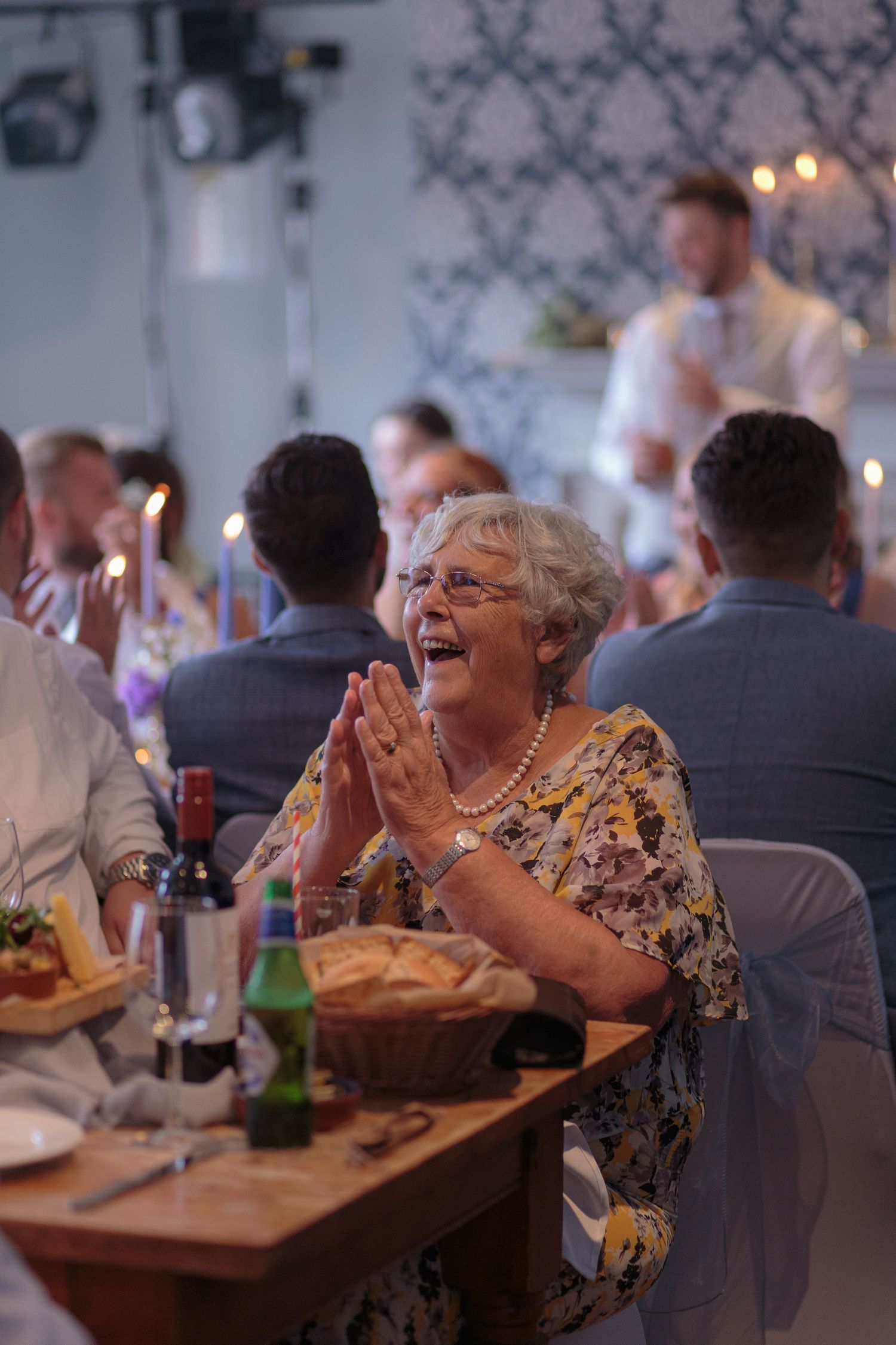 A woman sat laughing and clapping, as the groom gives his wedding speech behind her.