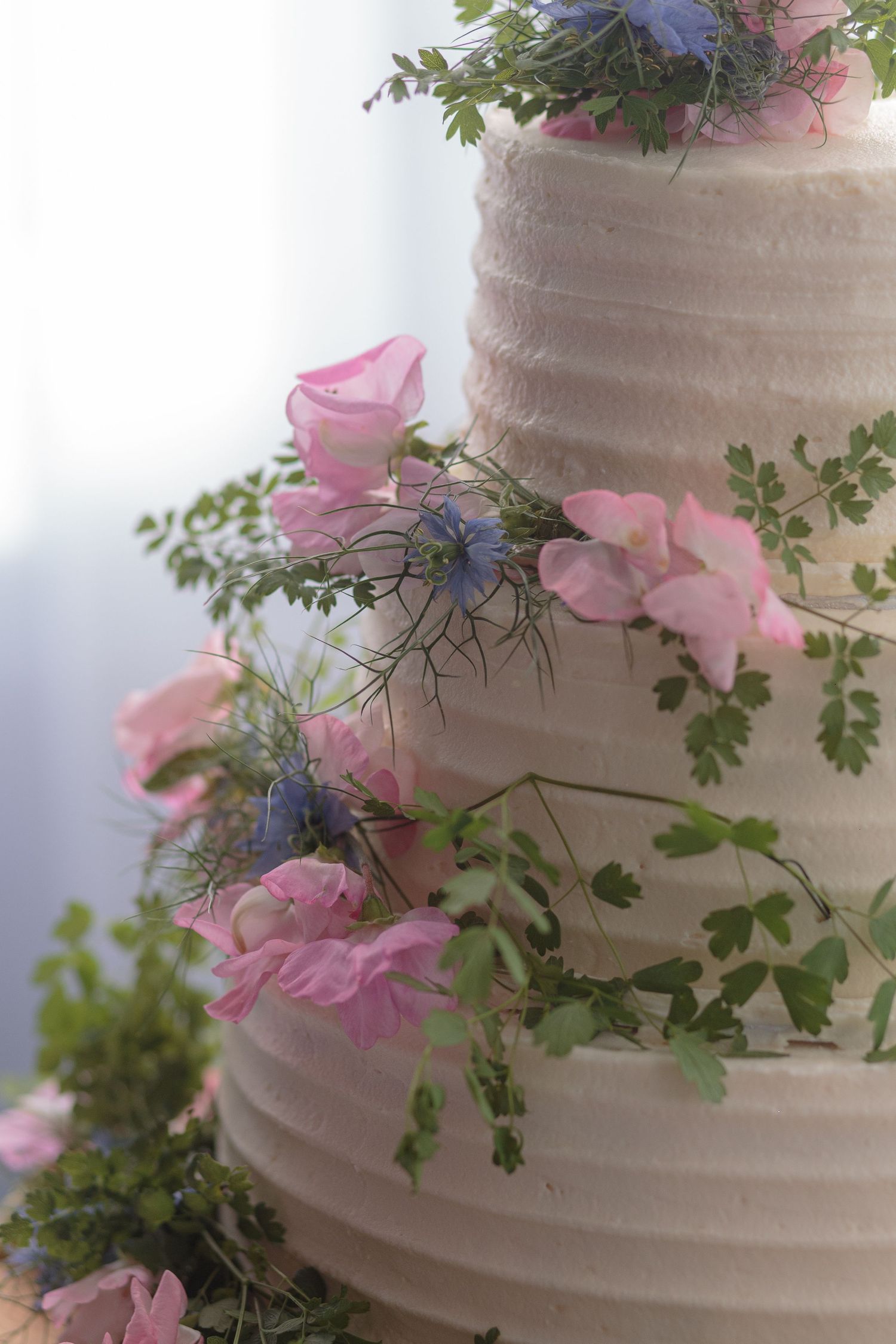 Close up details of pink and blue flowers, and lots of greenery, on the side of a beautiful white wedding cake.
