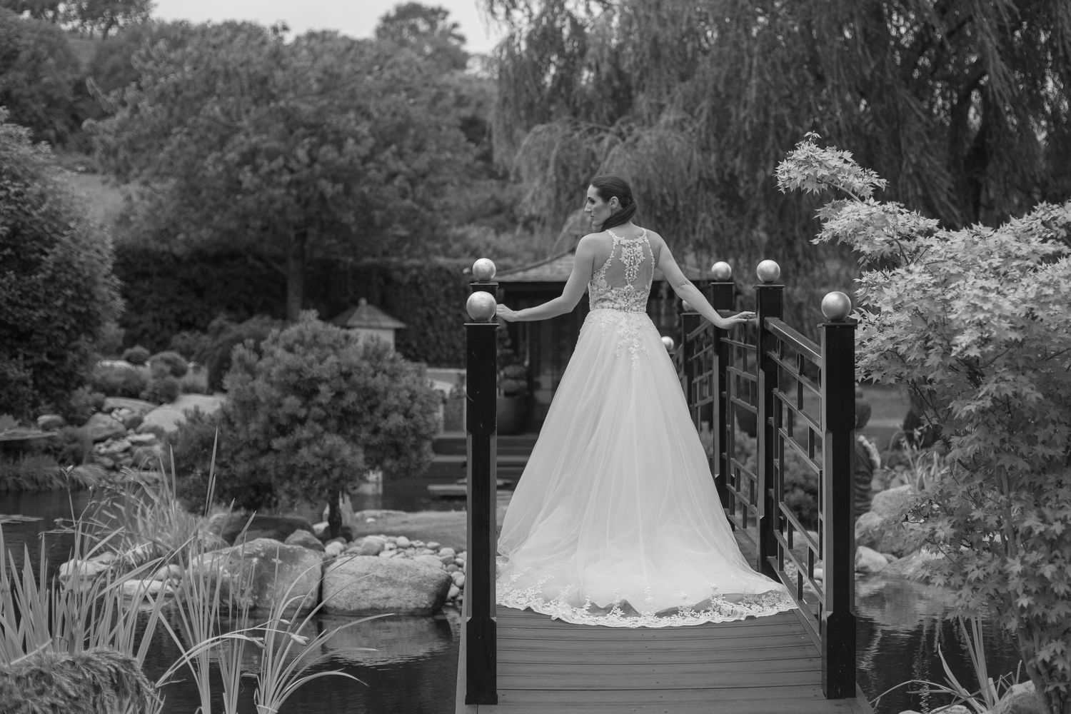 A bride walks across a bridge over a lake away from the camera, the skirt of her wedding dress billowing out behind her.