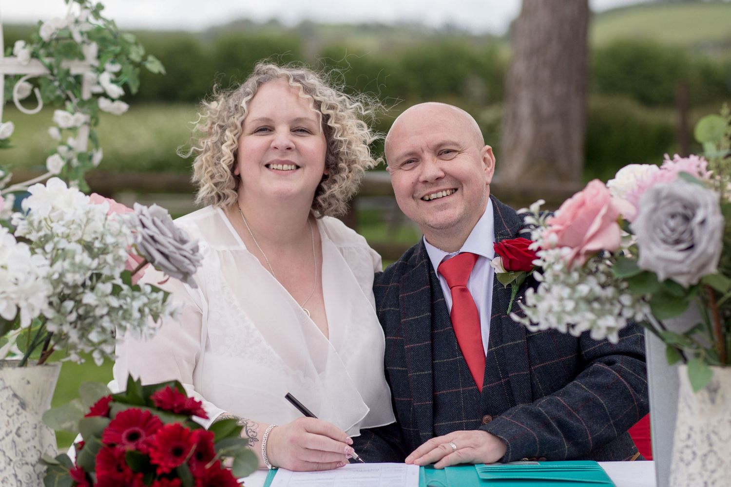 Bride and Groom smiling at the camera in the middle of signing their wedding license. Flowers sit in vases at the edges of their table.