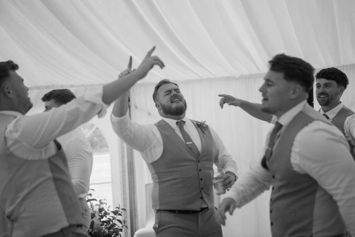 A group of groomsmen, hands raised in the air, dancing and singing to a favourite song on the dancefloor.