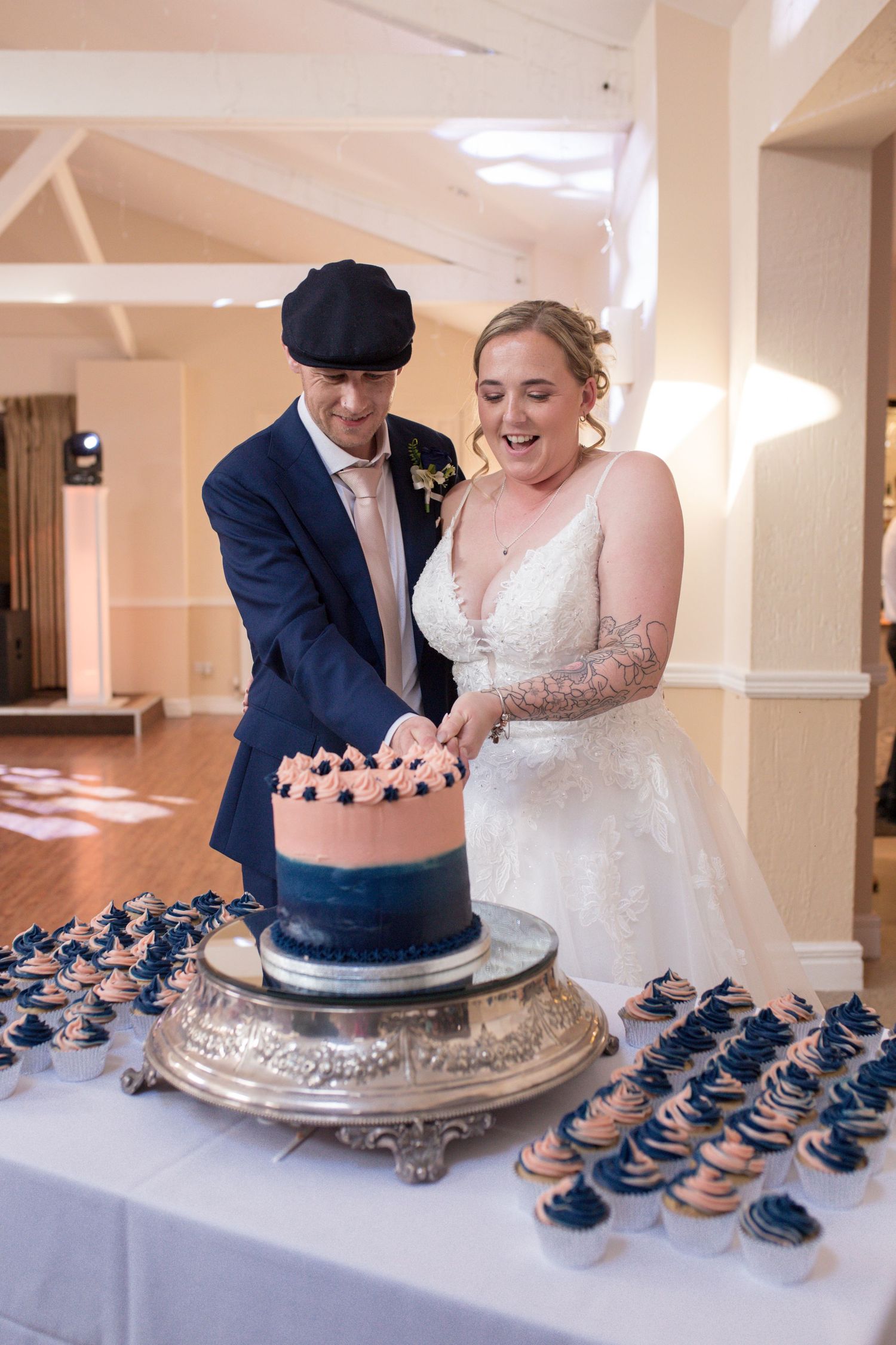 Bride and Groom cut into their pink and blue wedding cake together, surrounded by a table of matching cupcakes.