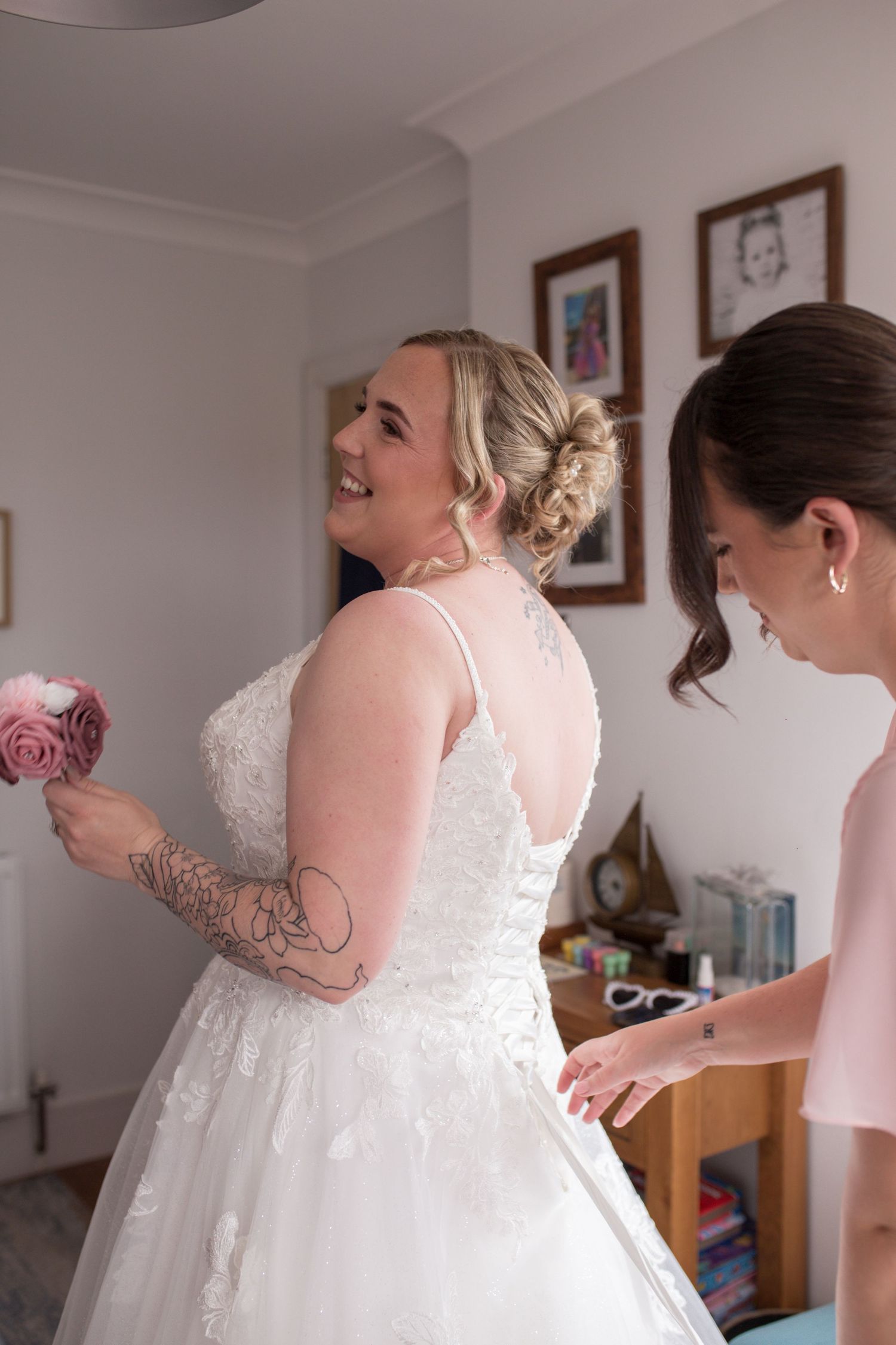 Bride smiles over her shoulder as one of her bridesmaids laces up the back of her wedding dress