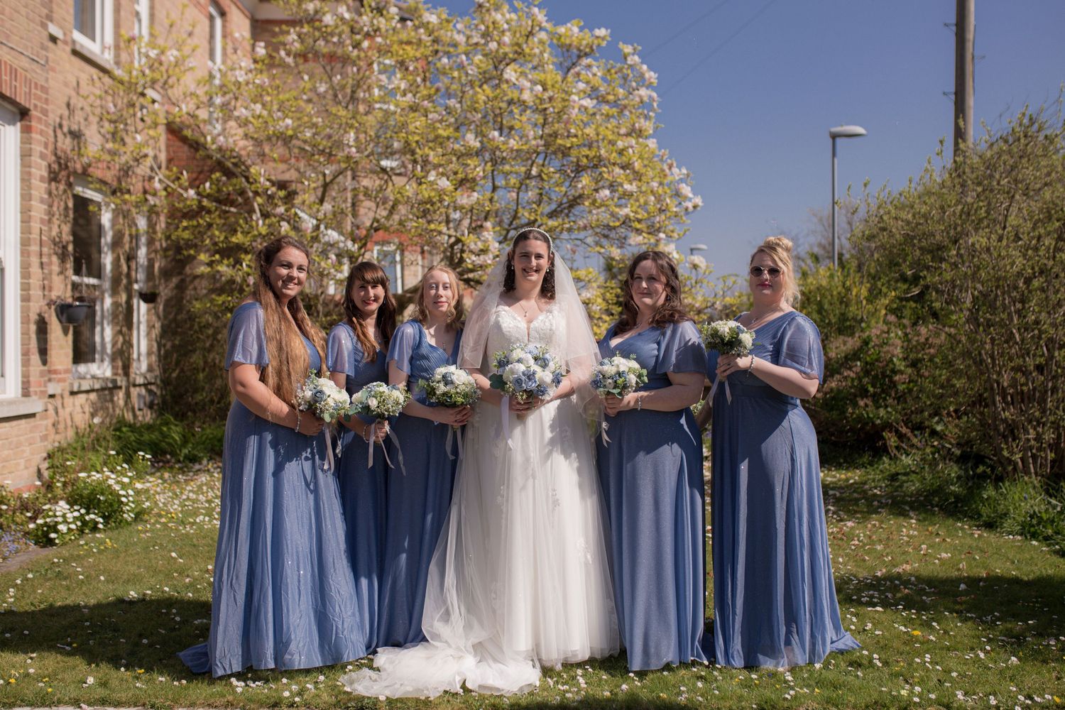 Bride and her five bridesmaids pose for a photo in the garden before the ceremony. Bridesmaids wearing blue, an d each holding a bouquet
