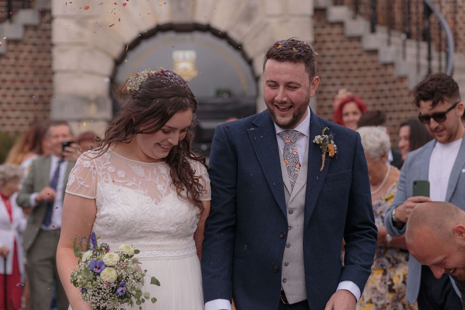 Bride and groom smile, covered in confetti and dried flower petals, their wedding guests surrounding them