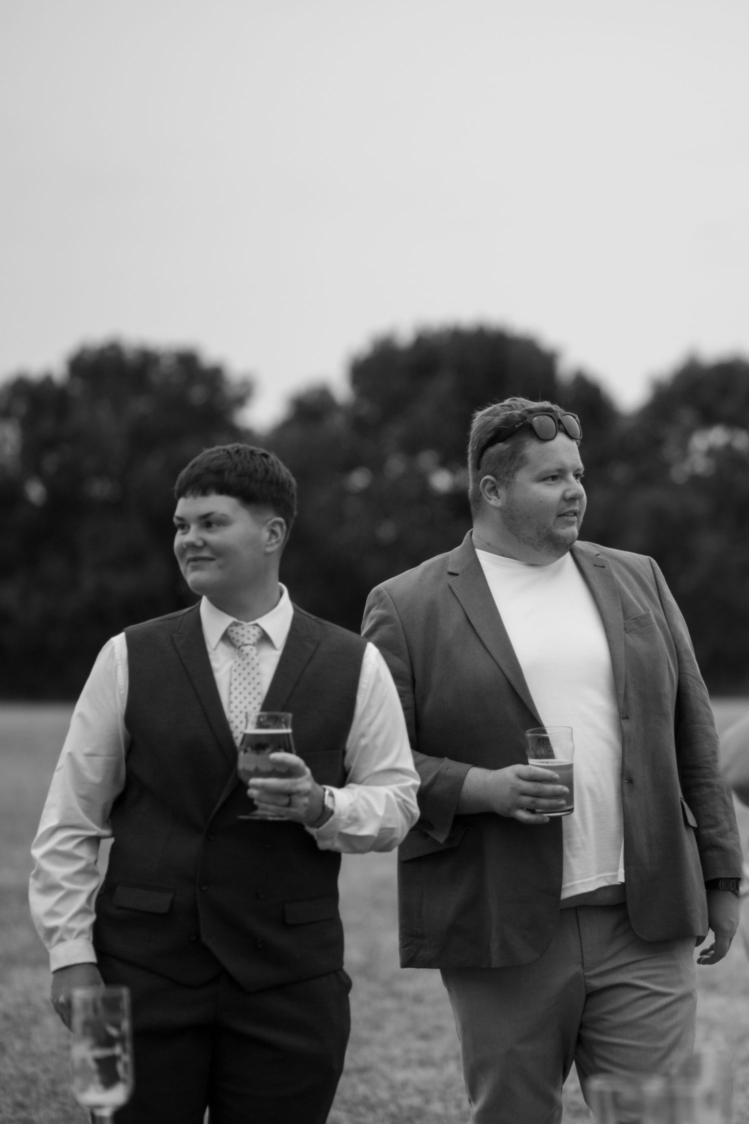 A black and white photo of two wedding guests stood together, drinks in hand, looking off in different directions.