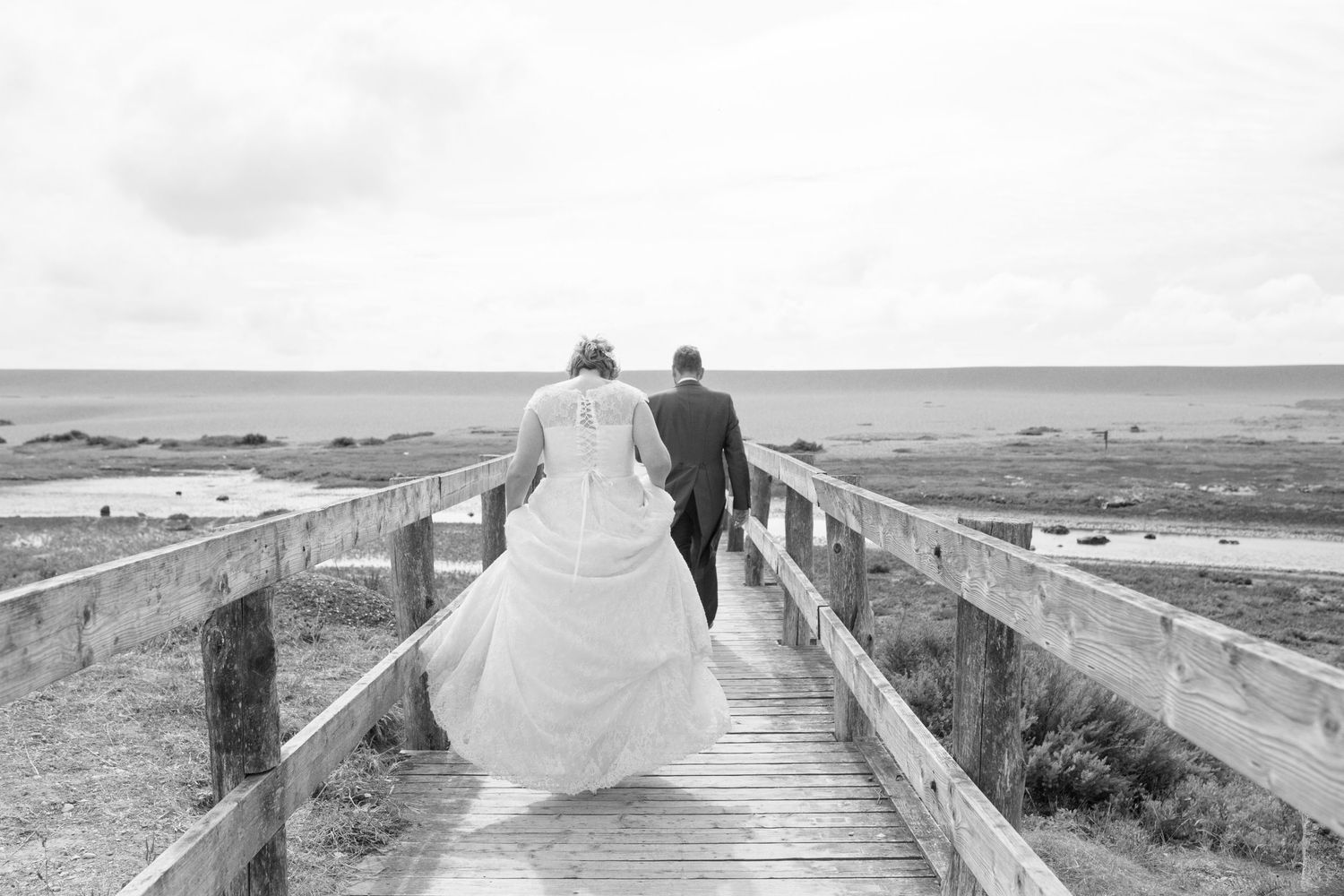A bride and groom walk across a wooden bridge heading onto Chesil Beach for portraits.