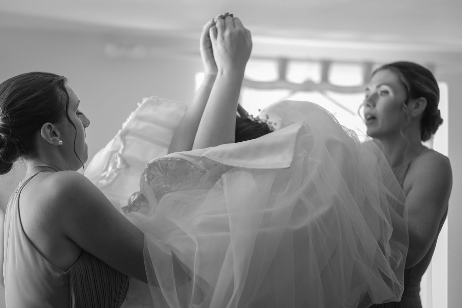 Black and White photos of three bridesmaids helping to get the wedding dress over the brides head