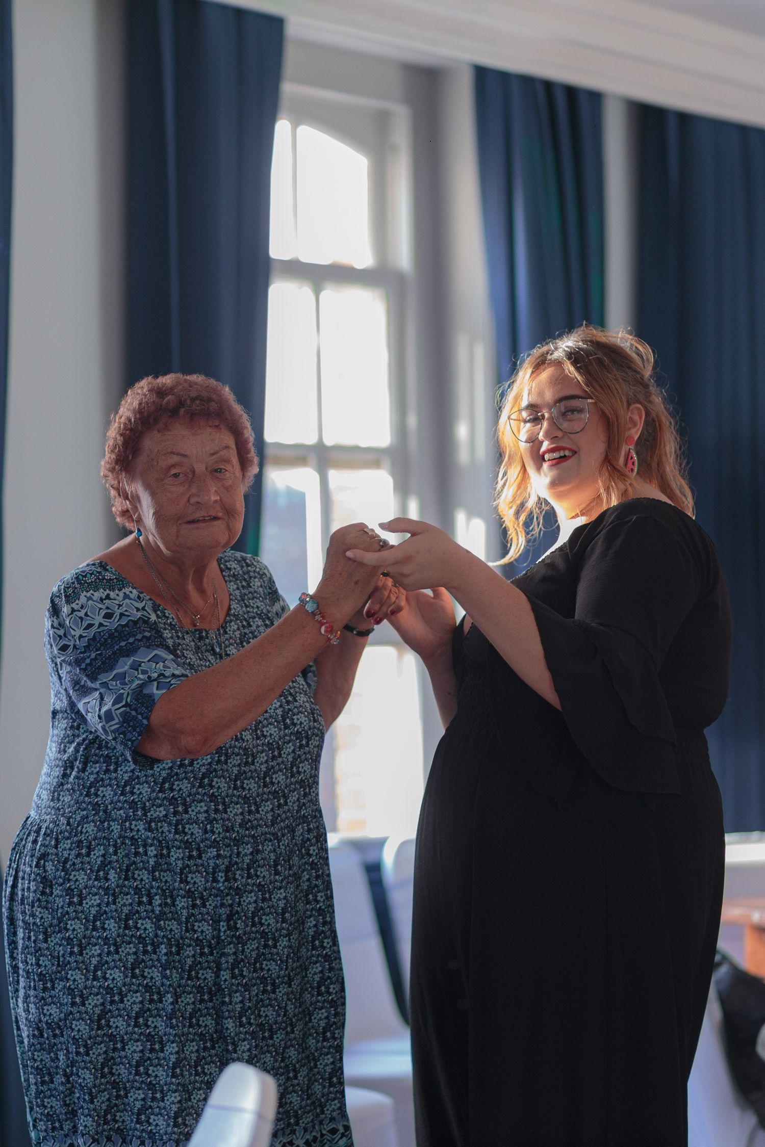 An older woman and her daughter smile for a photo, their hands held delicately between them as they dance.