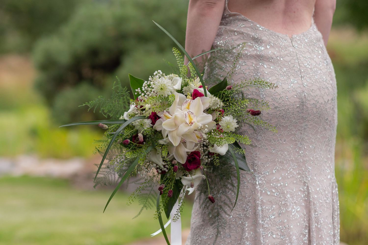 A bride faces away from the camera, her bouquet of white, red and green flowers facing towards the camera, resting against her hip.