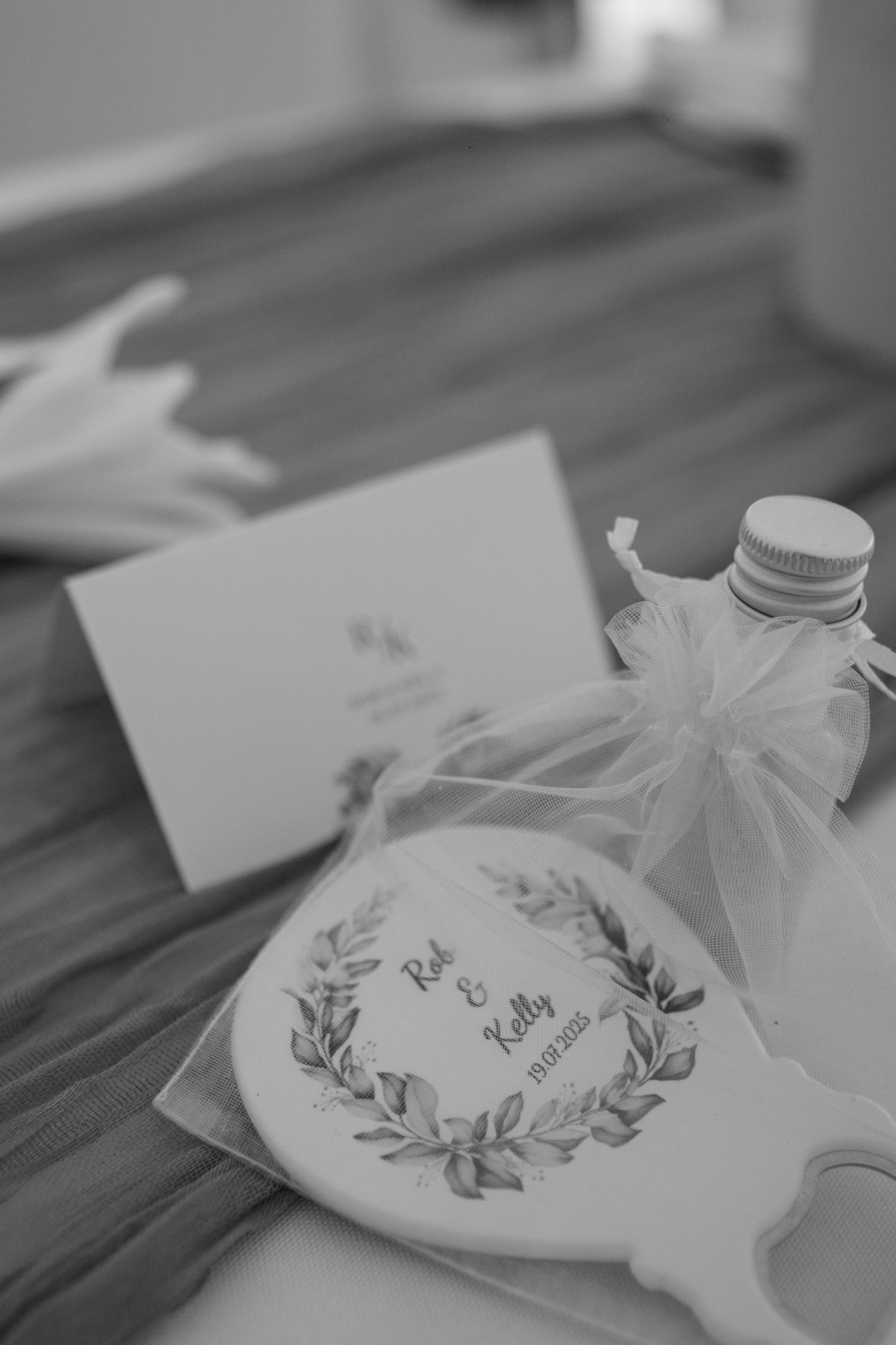 Black and white photo of wedding favours left on a table. A place card, a personalised bottle opener and a shot of alcohol.