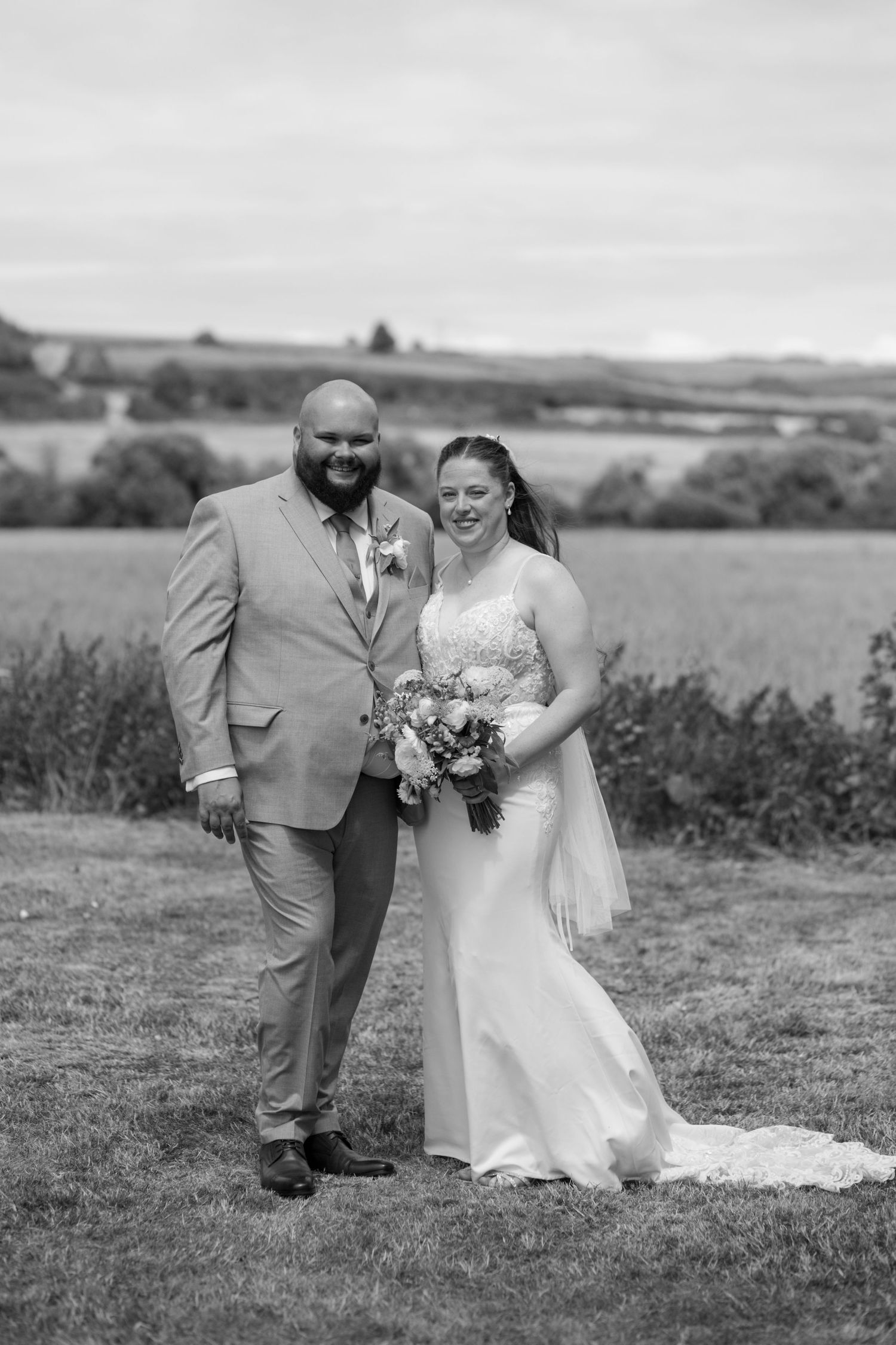 Black and white photo of a bride and groom stood together smiling surrounded by rolling hills and fields.