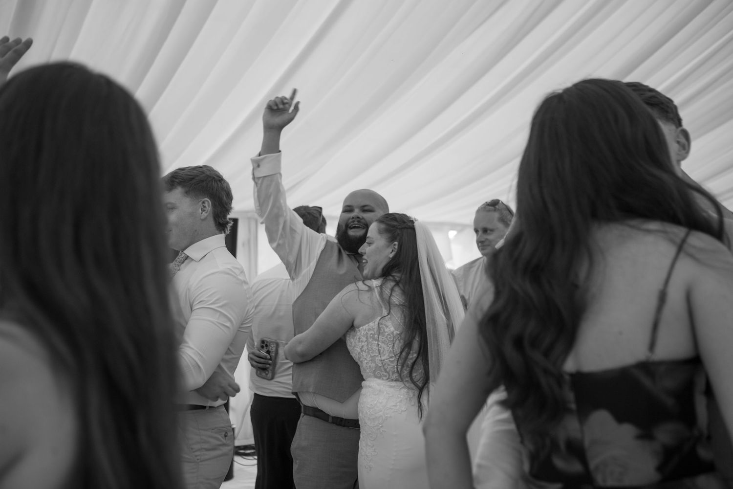 Bride hugging the Groom who has his arm stretched in the air, dancing together surrounded by their friends