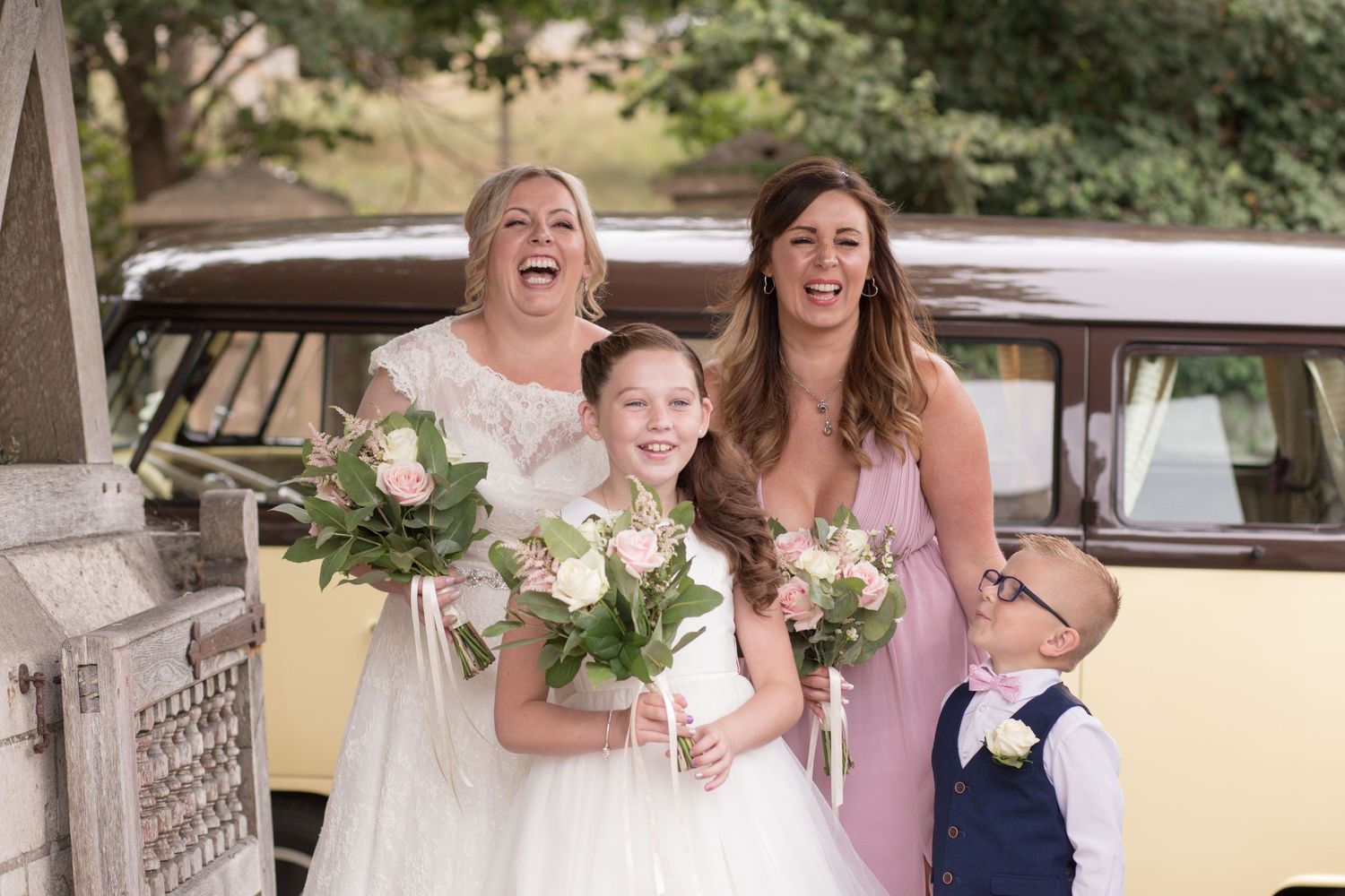 Bride laughs with her maid of honour, flower girl and page boy on their way into the church grounds