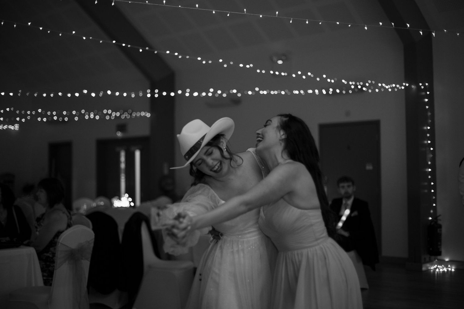 Bride wearing a cowboy hat, dances with her bridesmaid during the wedding reception, surrounded by glittering fairy lights