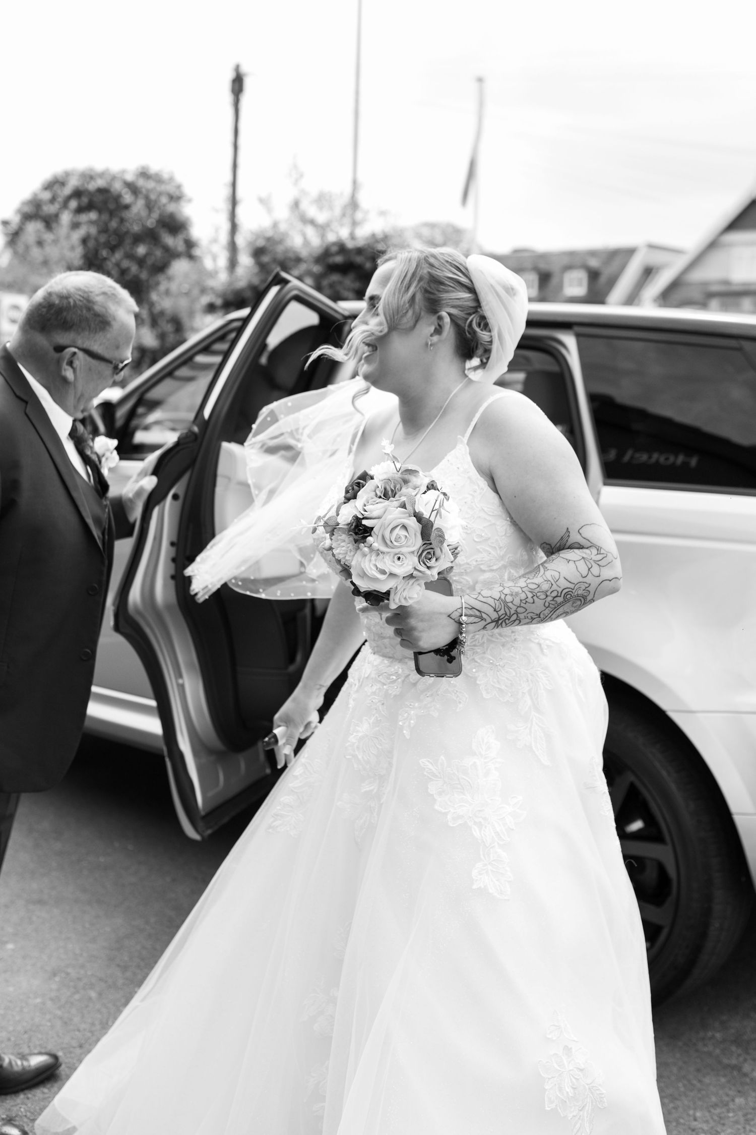 Black and White photo of a bride who has just stepped out of her wedding car. Her veil floating in the wind, her bouquet held proudly.