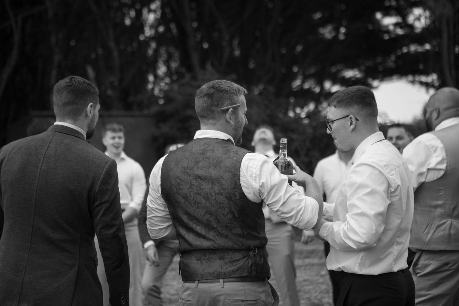 A group of men stood together in a circle passing a bottle of whiskey around them all. Shot from behind two men, as they pass the bottle.