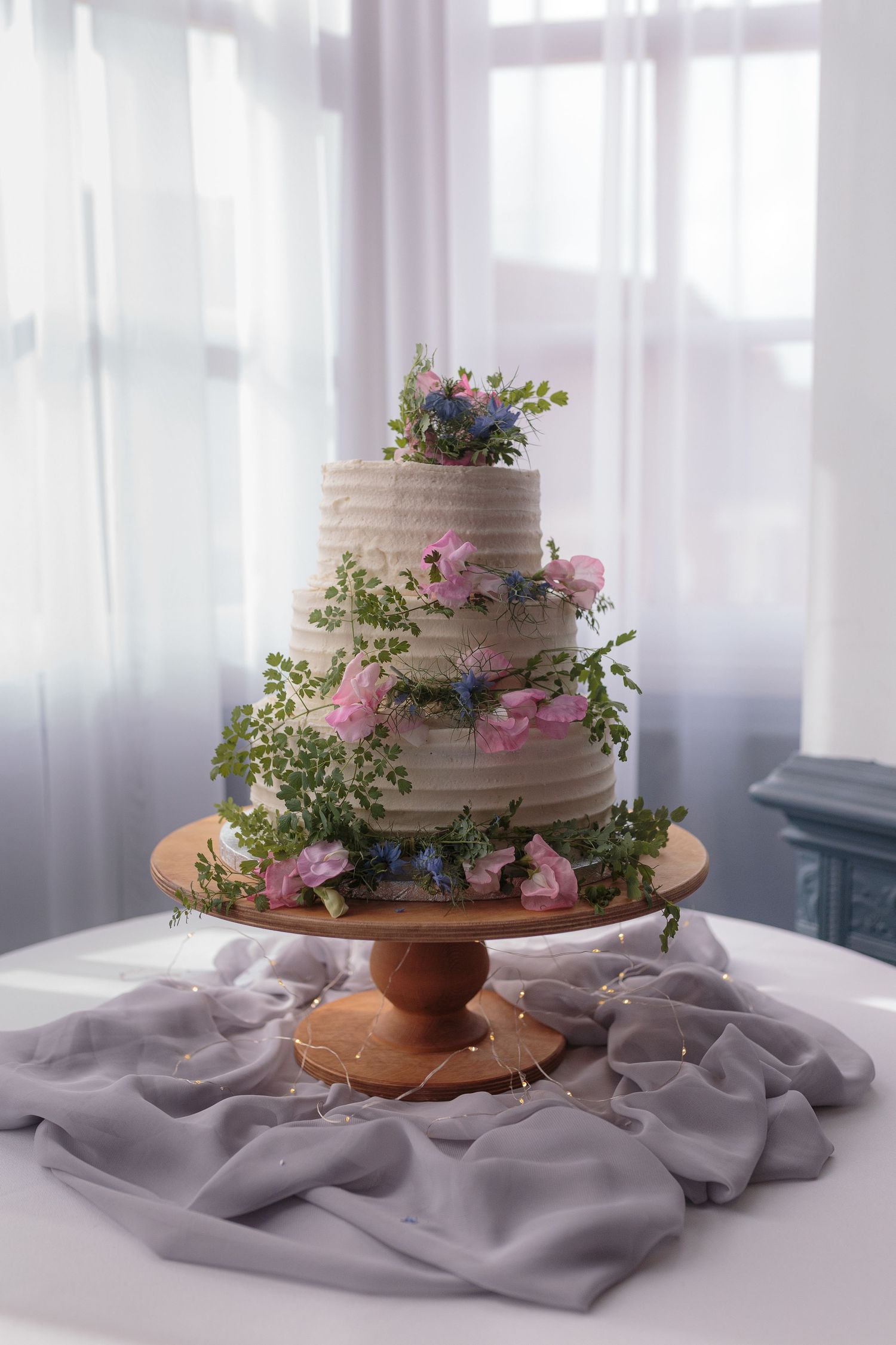 A white wedding cake, adjourned with pink and blue flowers and lots of greenery. Sits on a wooden cake stand on a table in the window