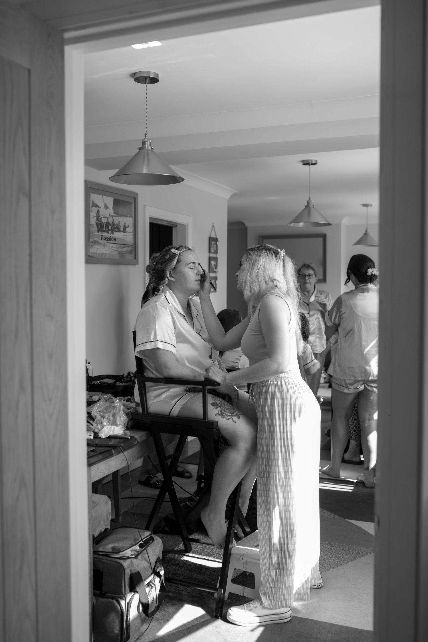 Black and White photo taken through a doorway, as a bride sits in a high chair having her make up done.
