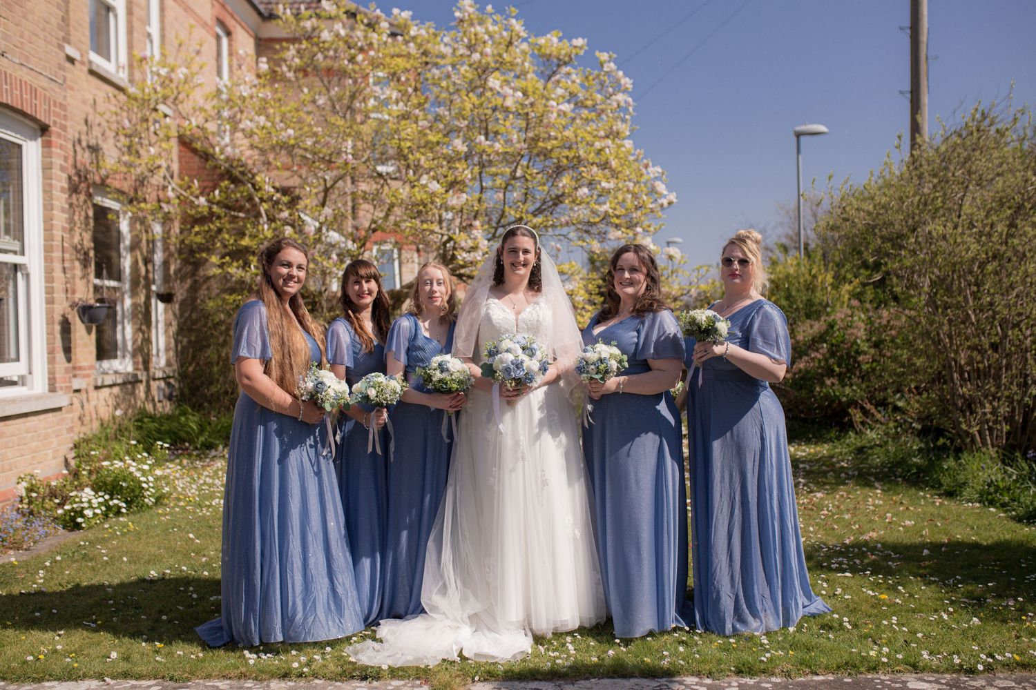 Bride and her five bridesmaids pose for a photo in the garden before the ceremony. Bridesmaids wearing blue, an d each holding a bouquet