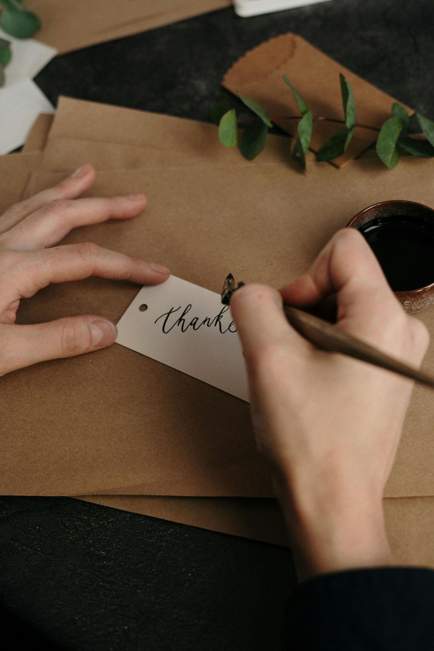 Hands writing 'thanks' on a gift tag with green foliage and brown paper in the background.