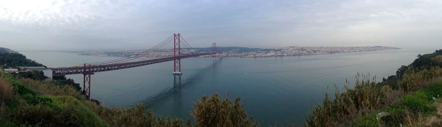 Wide panoramic view of the 25 de Abril Bridge spanning Lisbon's Tagus River.