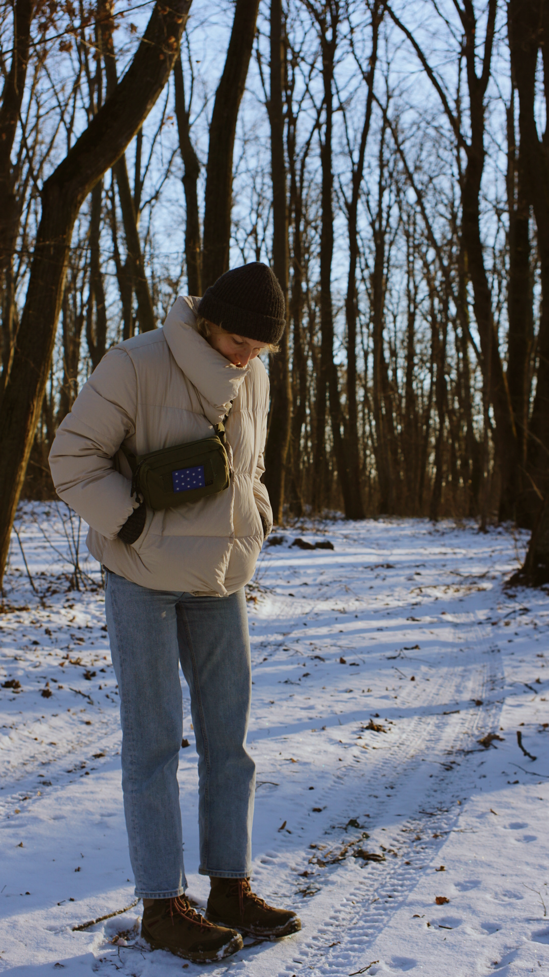 A person in winter attire with a small crossbody bag stands on a snow-covered forest trail.