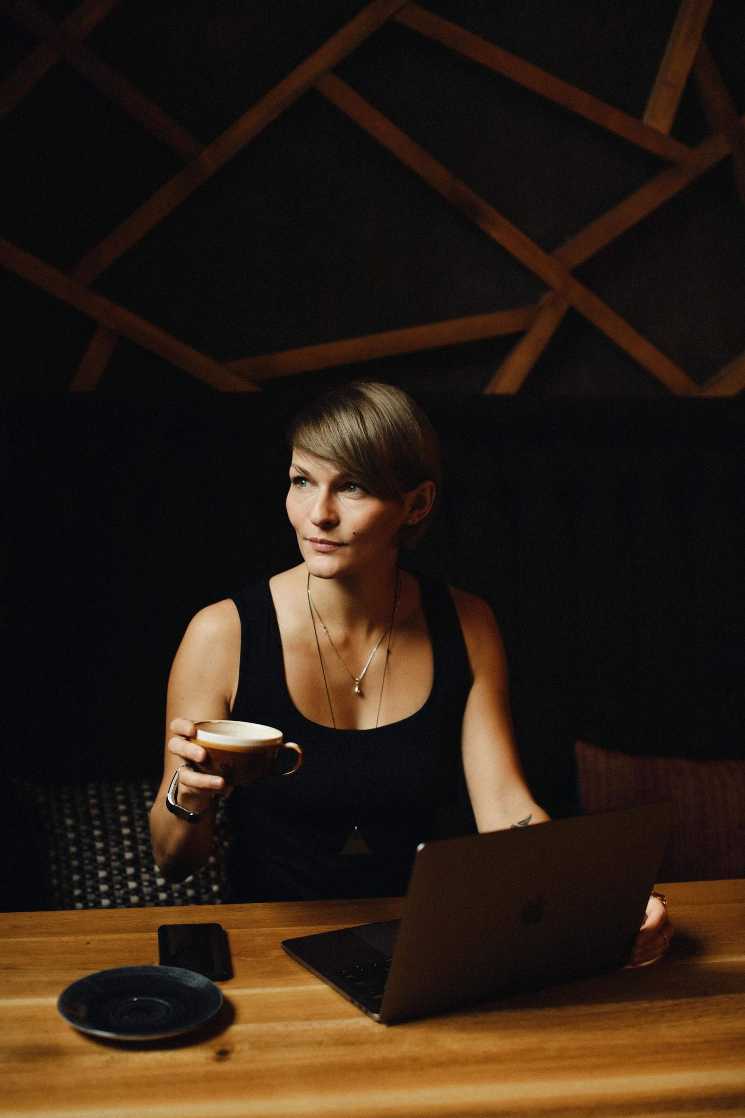 Person sitting at wooden table with laptop and coffee in dark room with geometric wall patterns.