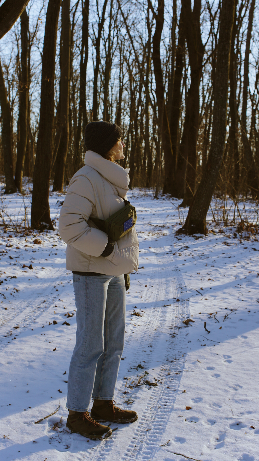Person in a cream puffer coat and gray pants walking through a snowy winter forest trail.