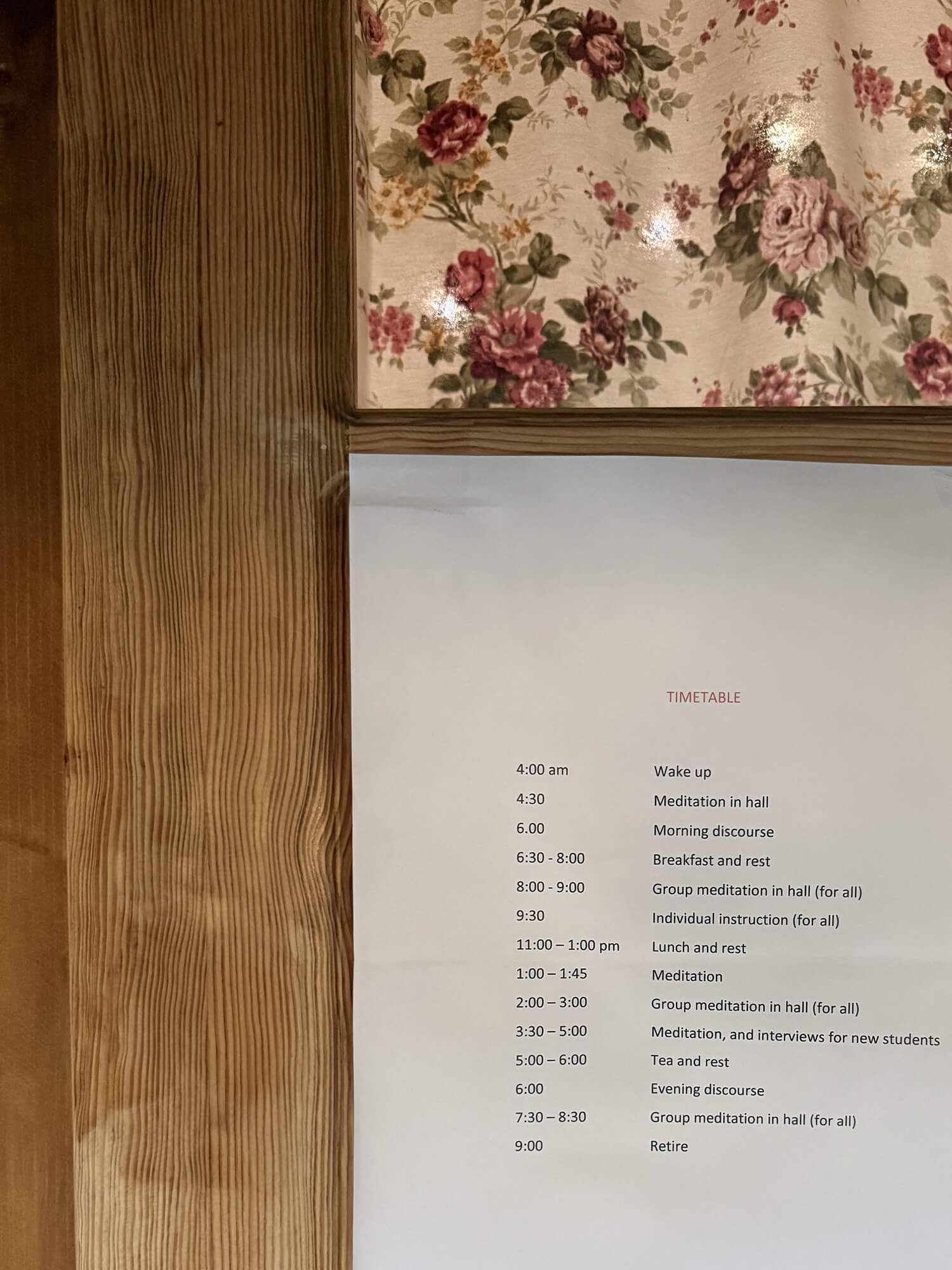 A timetable schedule posted on a wooden wall beside floral fabric showing daily meditation retreat activities.