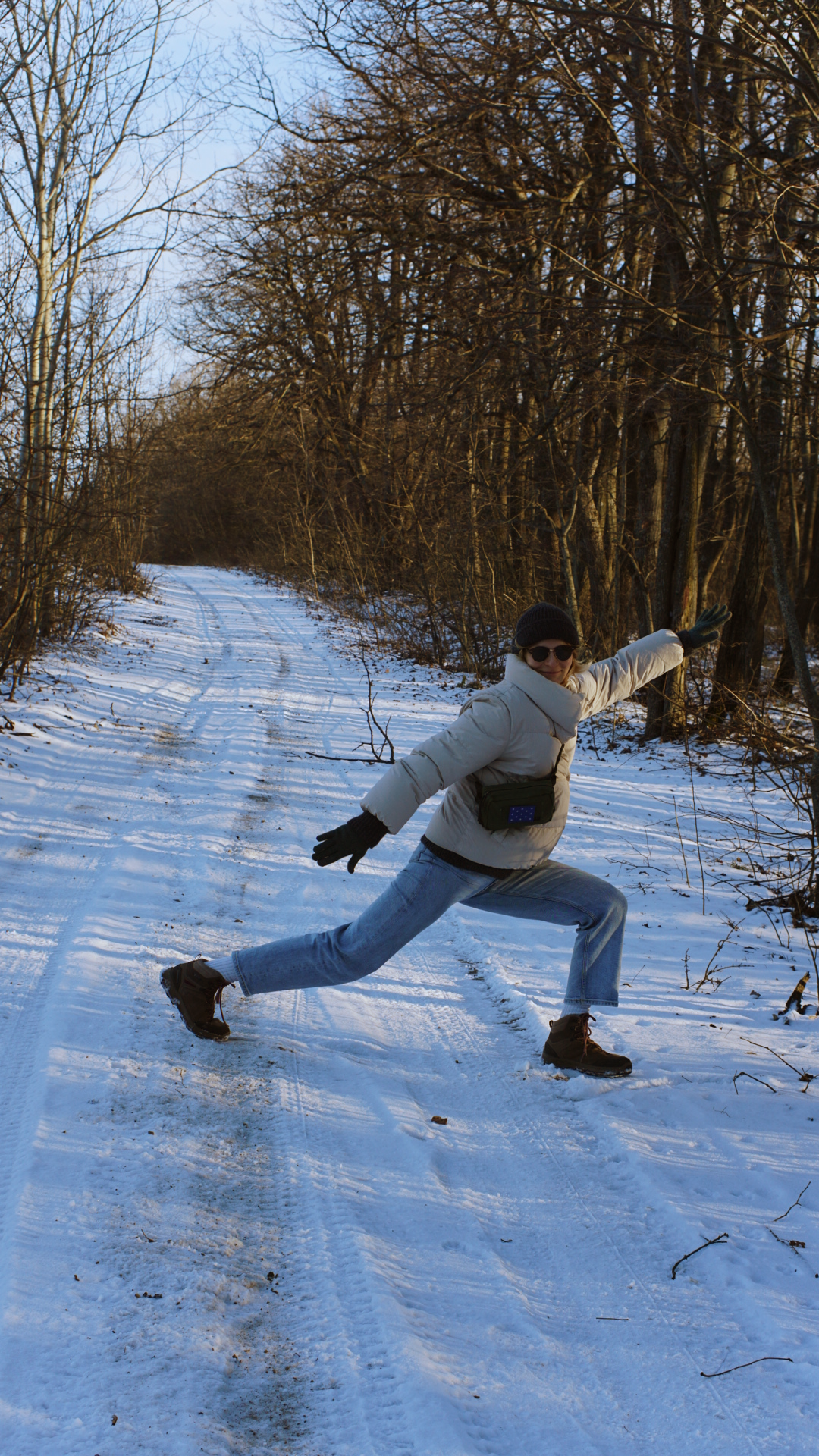 A playful winter walker strikes a dramatic pose on a snowy forest trail while wearing a light jacket.