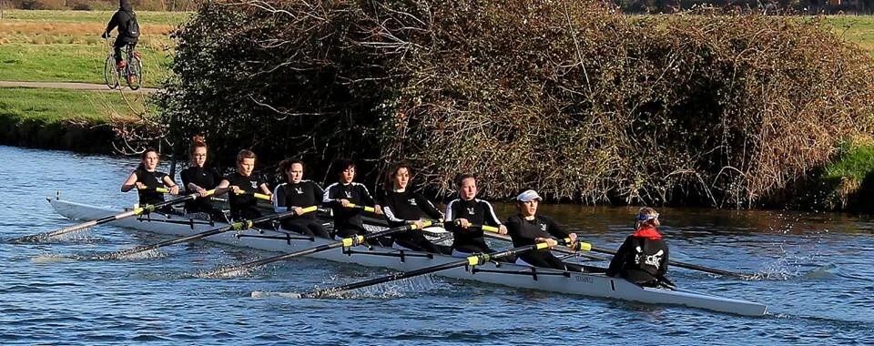 Rowing team practicing on water with multiple boats visible in sequence.