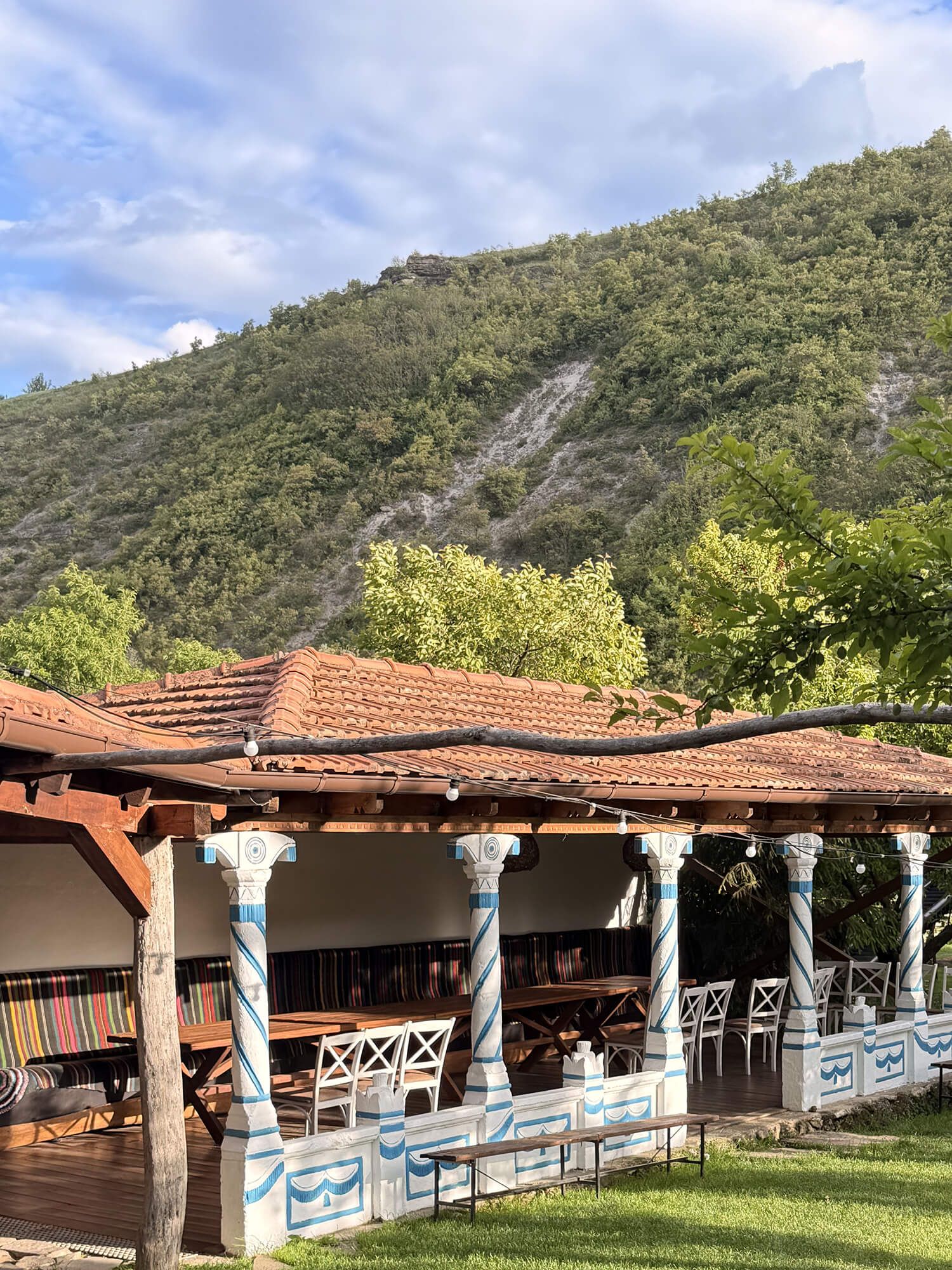 Rustic open-air pavilion with terracotta roof and decorated columns set against a lush green mountain backdrop.