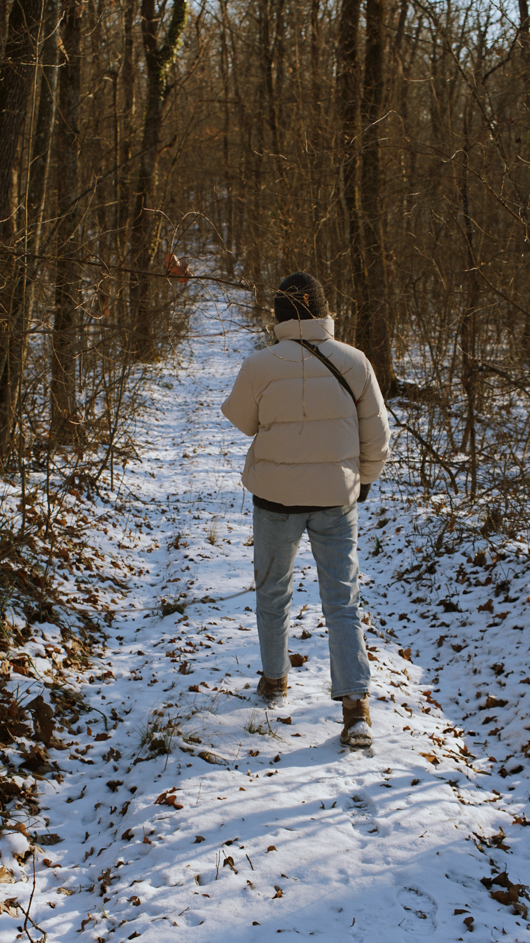Person in a cream puffer jacket and jeans walks through a snow-covered forest trail.