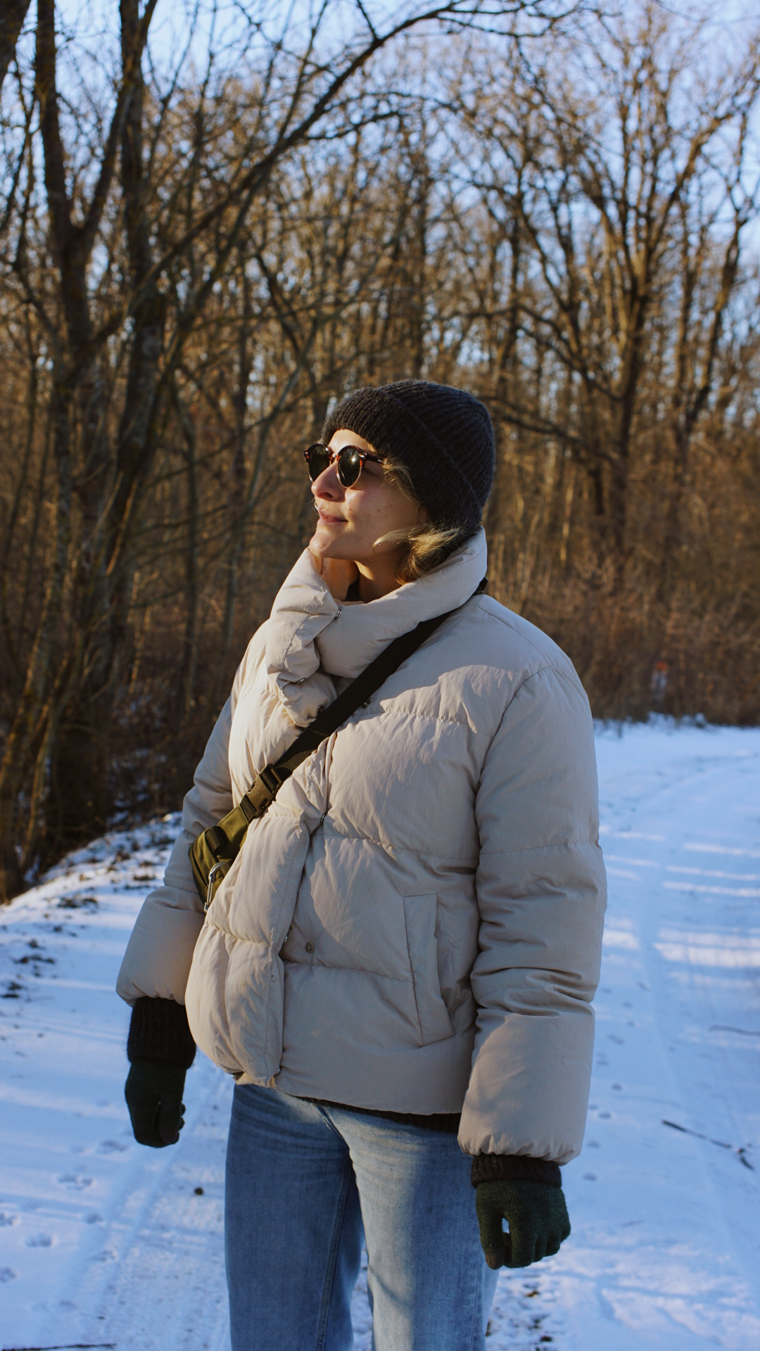 A winter walker in a cream puffer coat and beanie enjoys a snowy forest path on a sunny day.