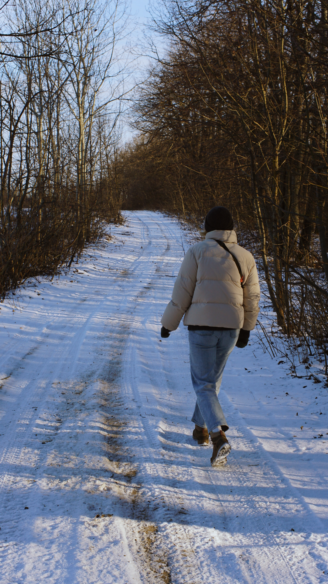 A solitary figure walks down a snowy forest path wearing a light-colored winter jacket and jeans.