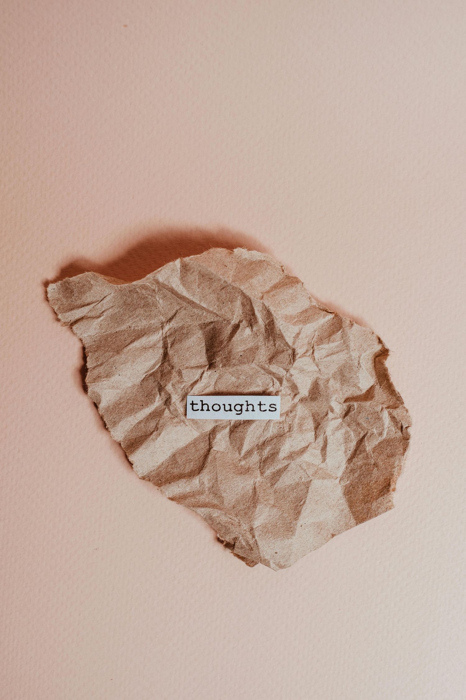 Crumpled brown paper with the word 'thoughts' on a pale pink background.